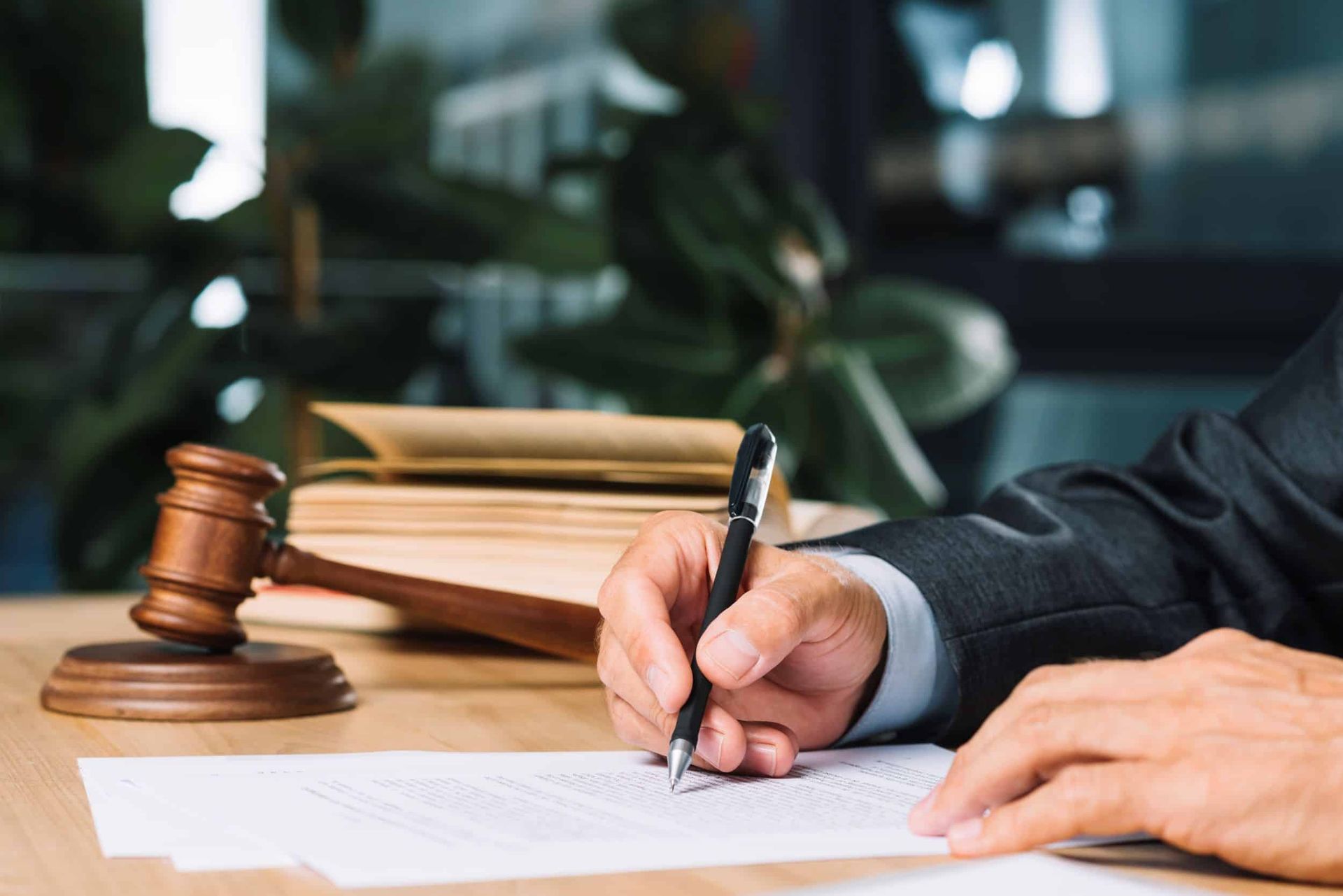 A person in a suit signing a document at a desk, gavel and books in the background.