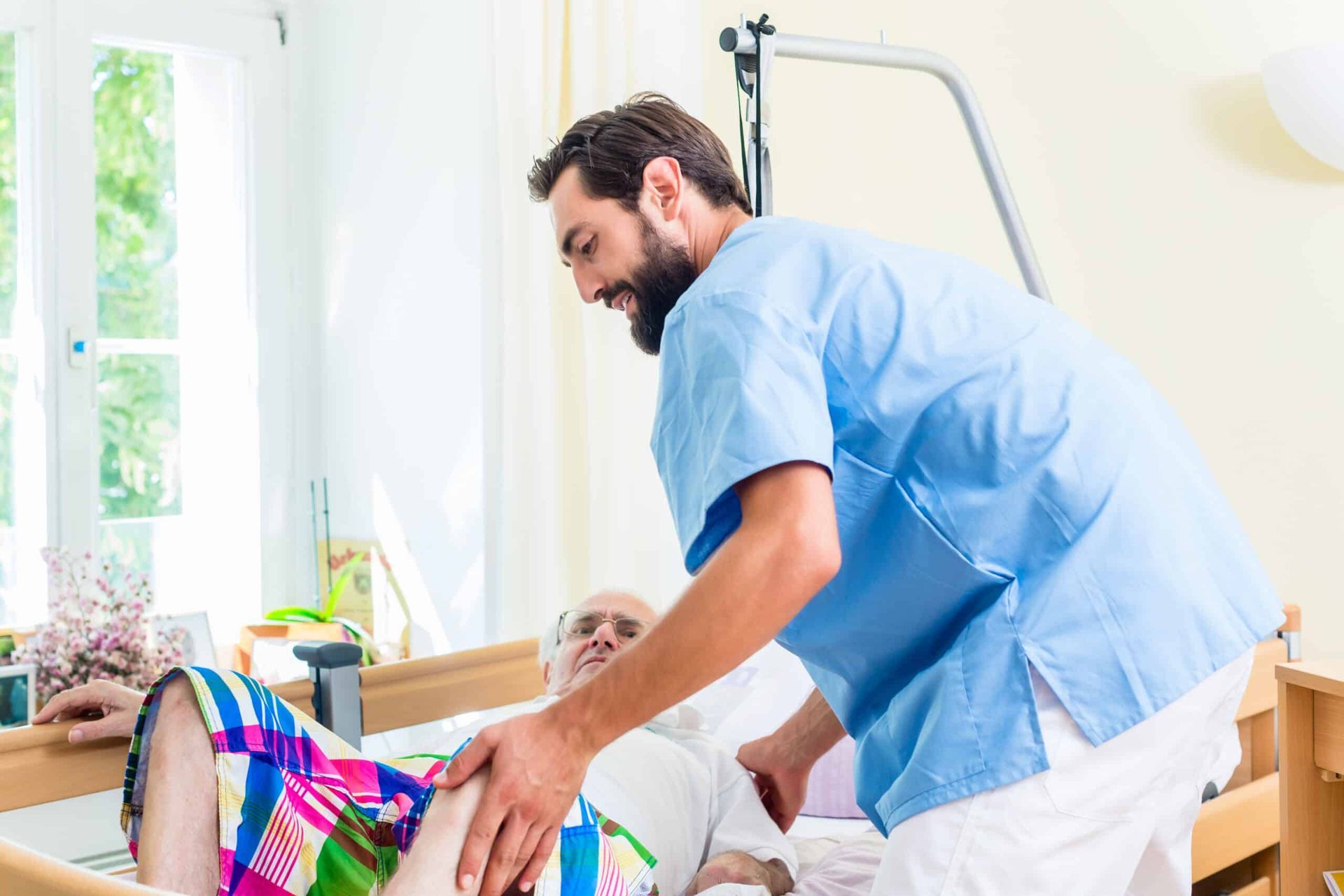 Caregiver assists elderly patient out of bed; light-blue scrubs in a bedroom setting.