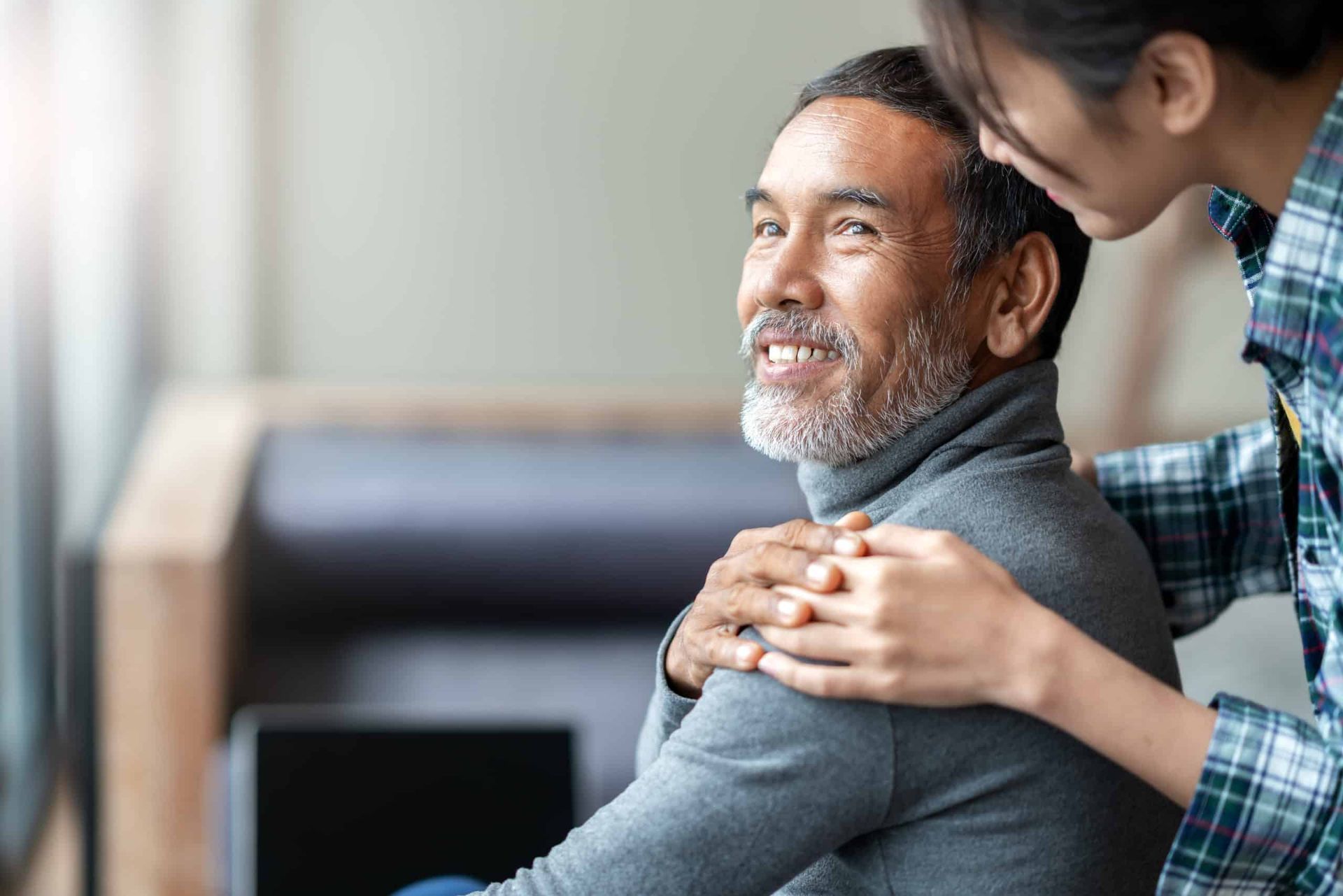 Smiling older man with gray beard, embraced by a younger woman, indoors.