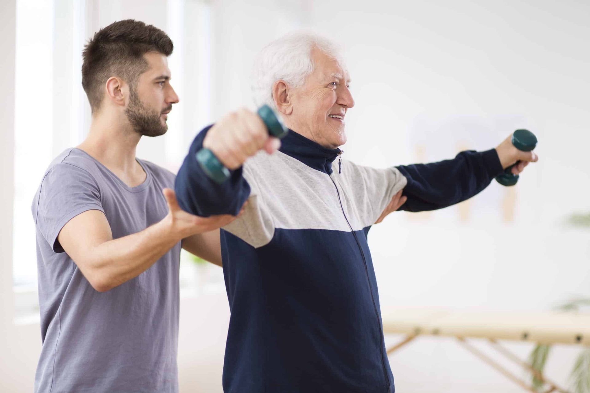 A young man assists an elderly man lifting small weights, exercising indoors.