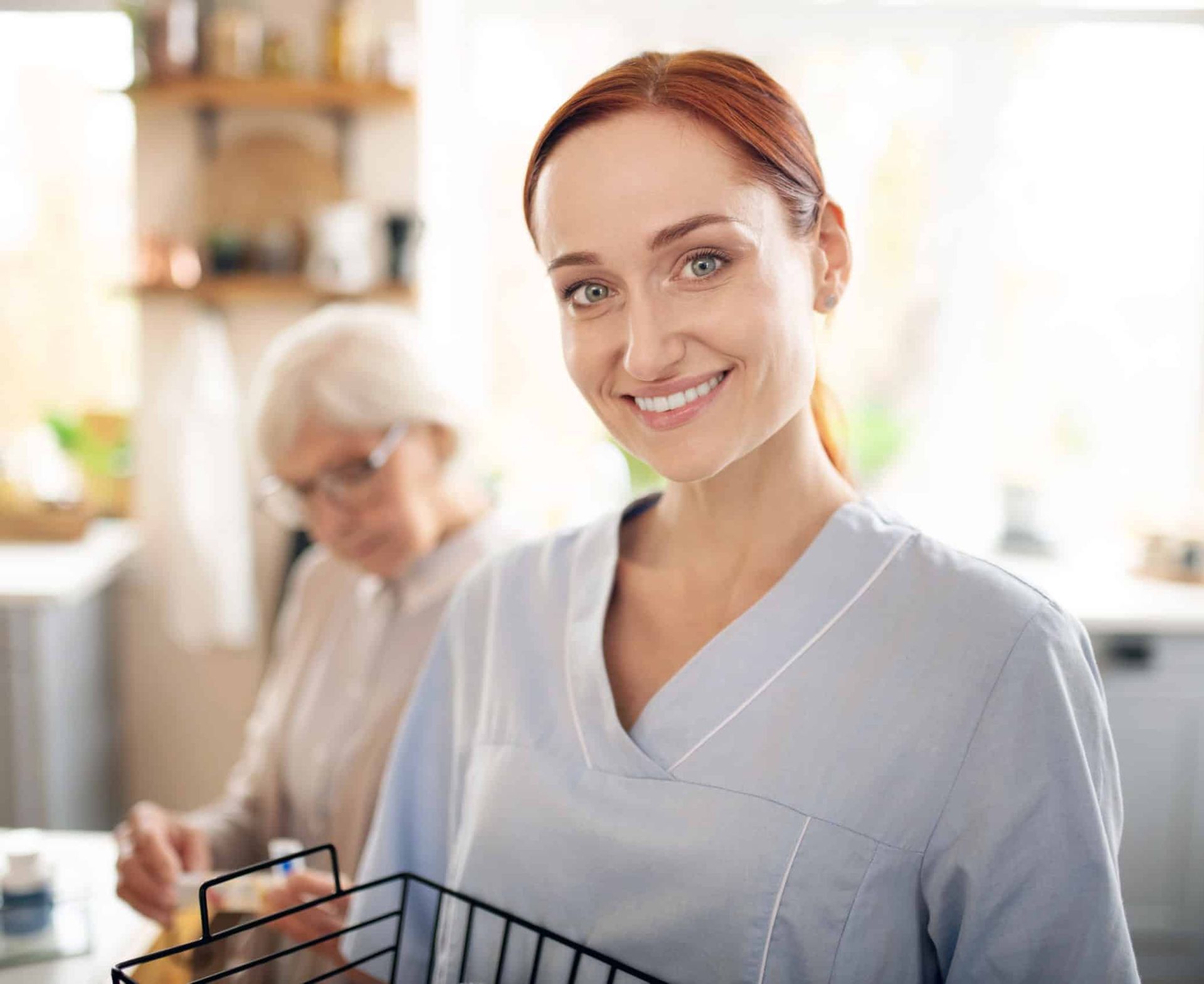Caregiver smiles, holding a basket, while assisting an older woman in a kitchen.