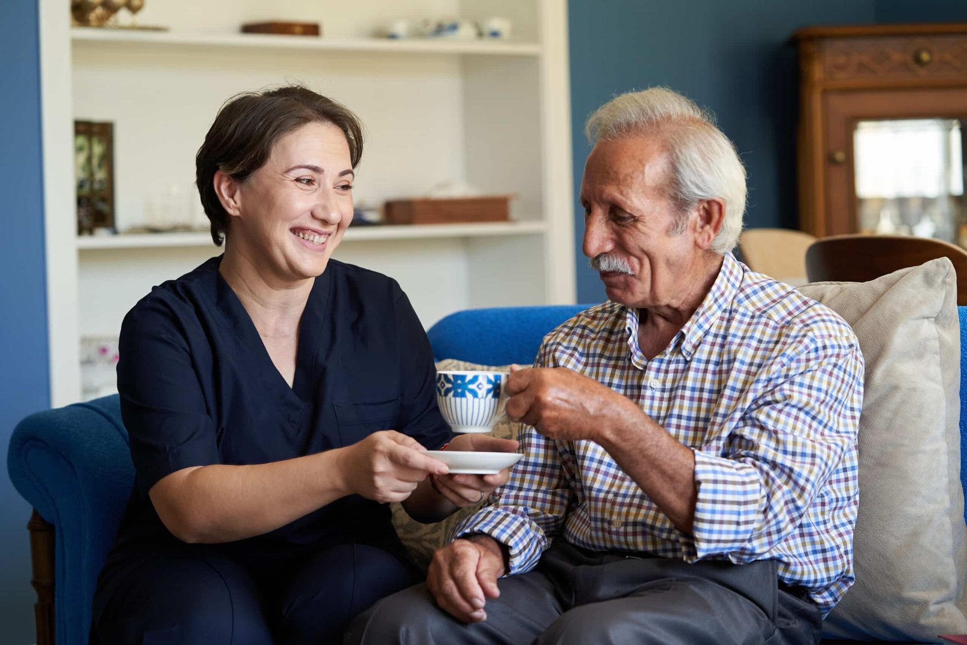 Caregiver smiles, offering tea to an elderly man on a blue sofa. They sit indoors with a bookcase.