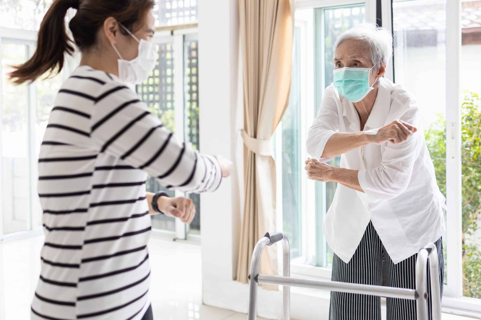 Woman in stripes and elderly woman with walker, both in masks, bumping elbows near window.