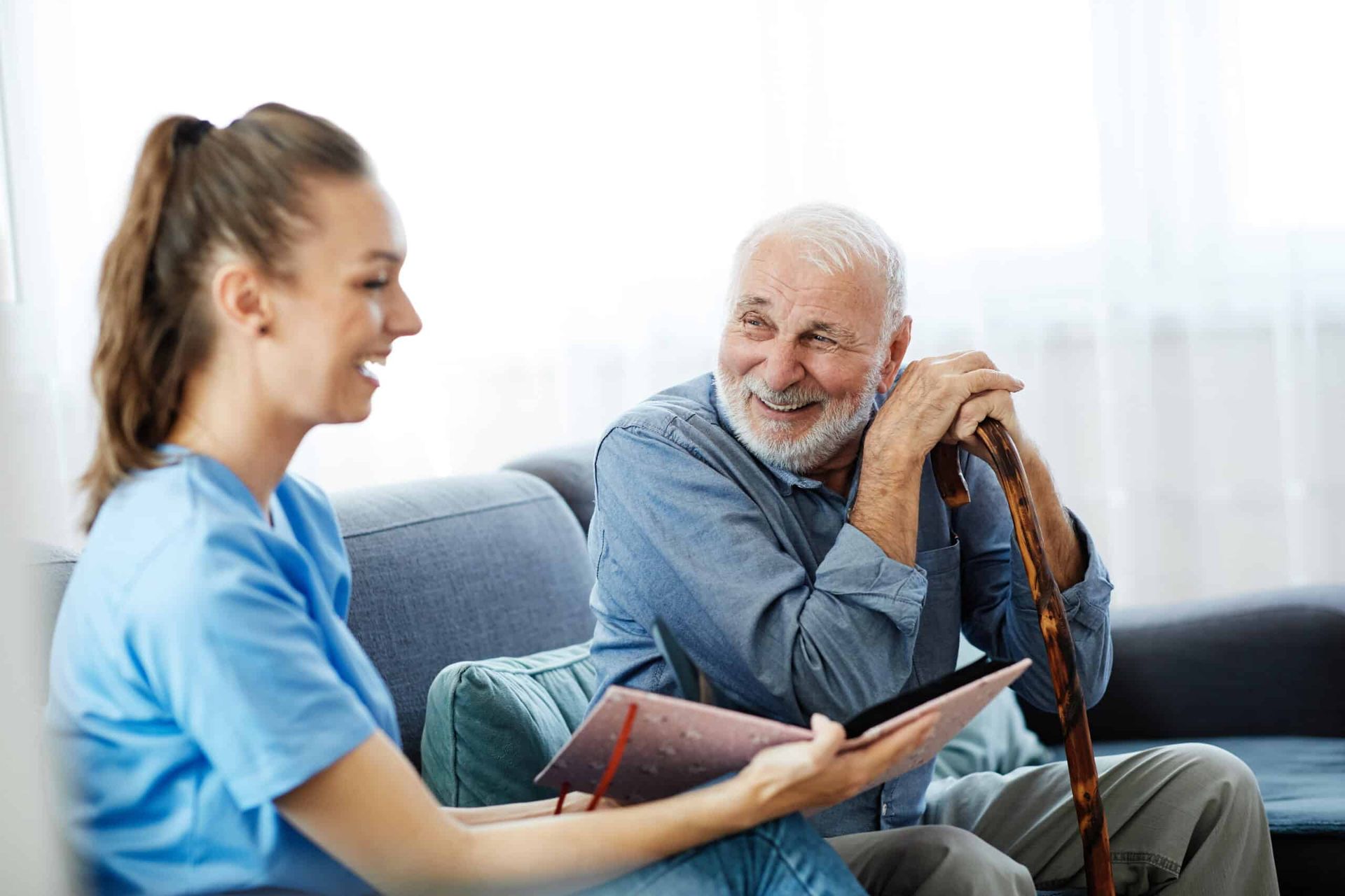 Young female caregiver and elderly man on a couch looking at a tablet, smiling.