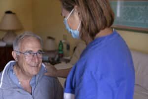 Nurse in blue scrubs, with face mask, comforts elderly man in a bedroom.