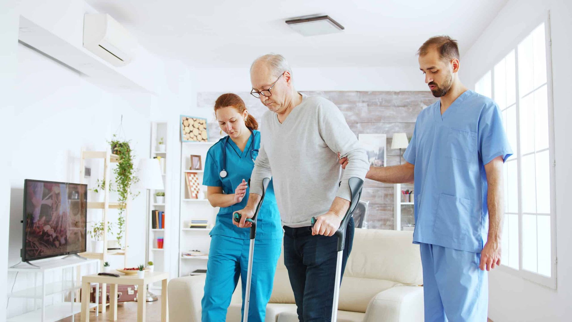 Elderly man using crutches with support from two medical professionals indoors.