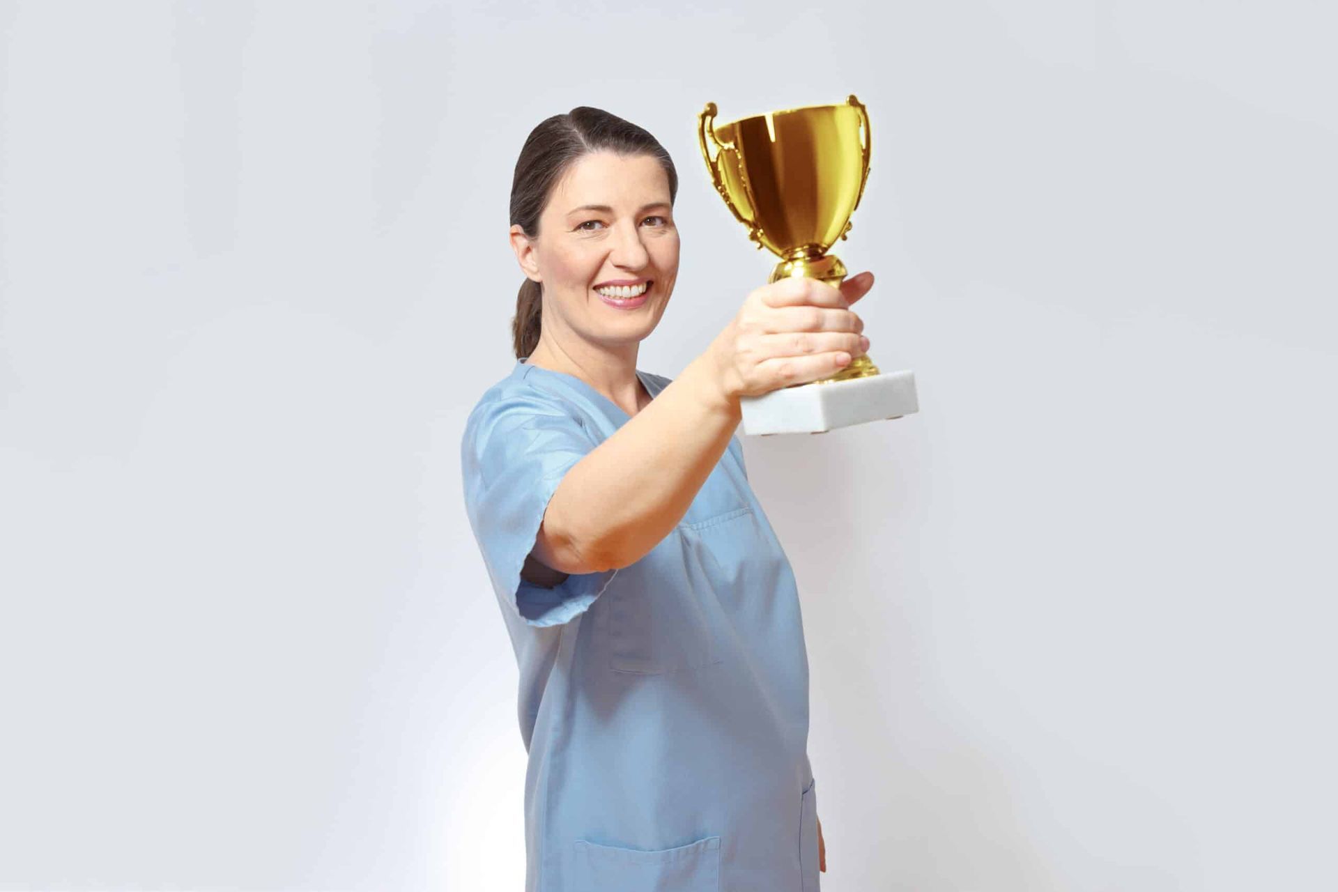 Woman in blue scrubs holds up a gold trophy, smiling in front of a white background.
