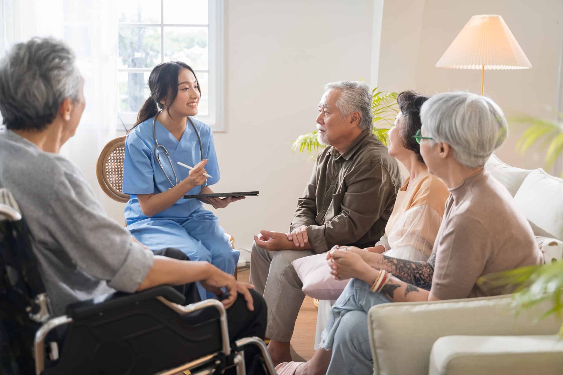 Nurse in blue scrubs with a tablet speaks to a group of elderly patients in a living room.