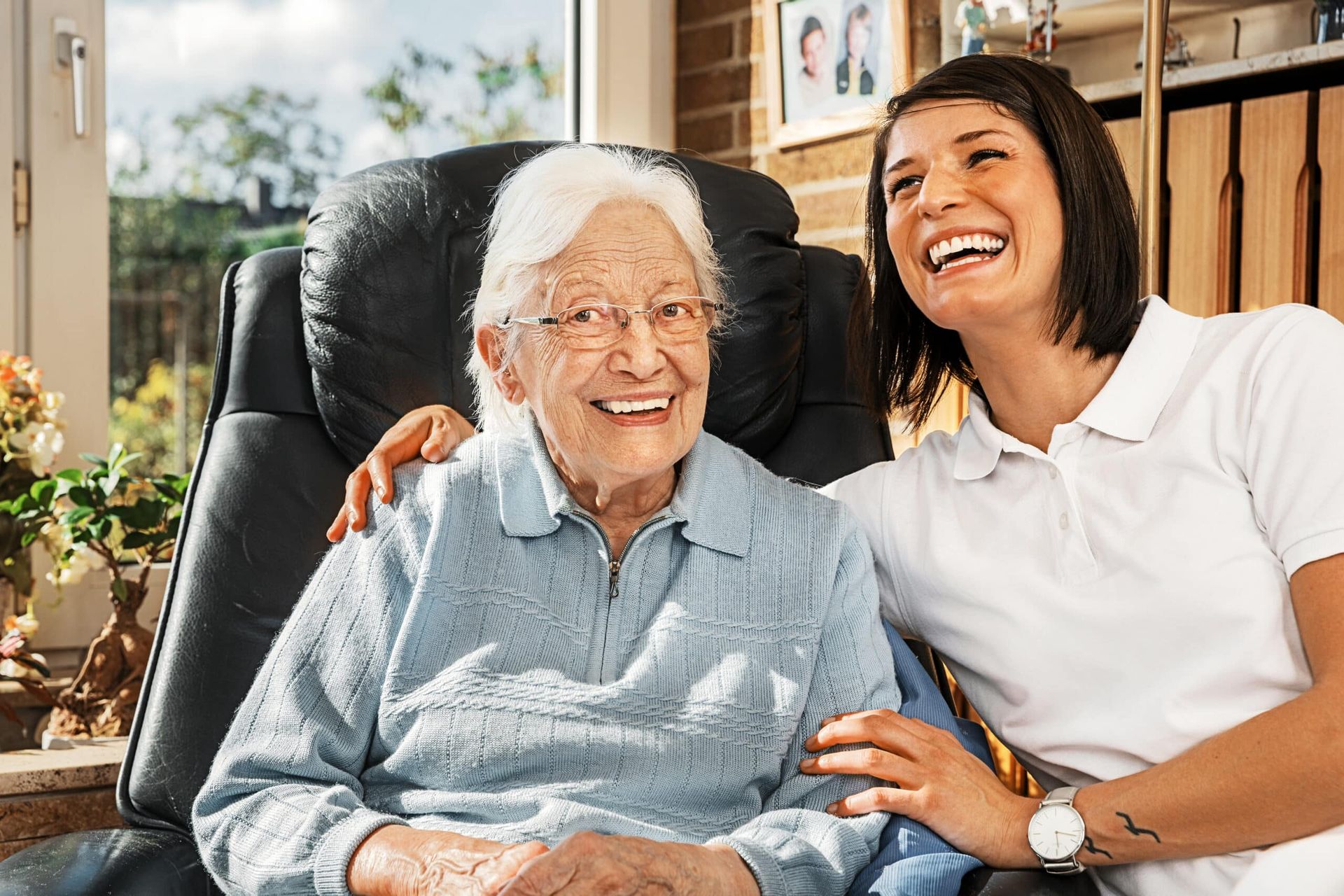 Elderly woman and caregiver smiling together. Woman in armchair, light blue top, caregiver in white, warm indoor setting.