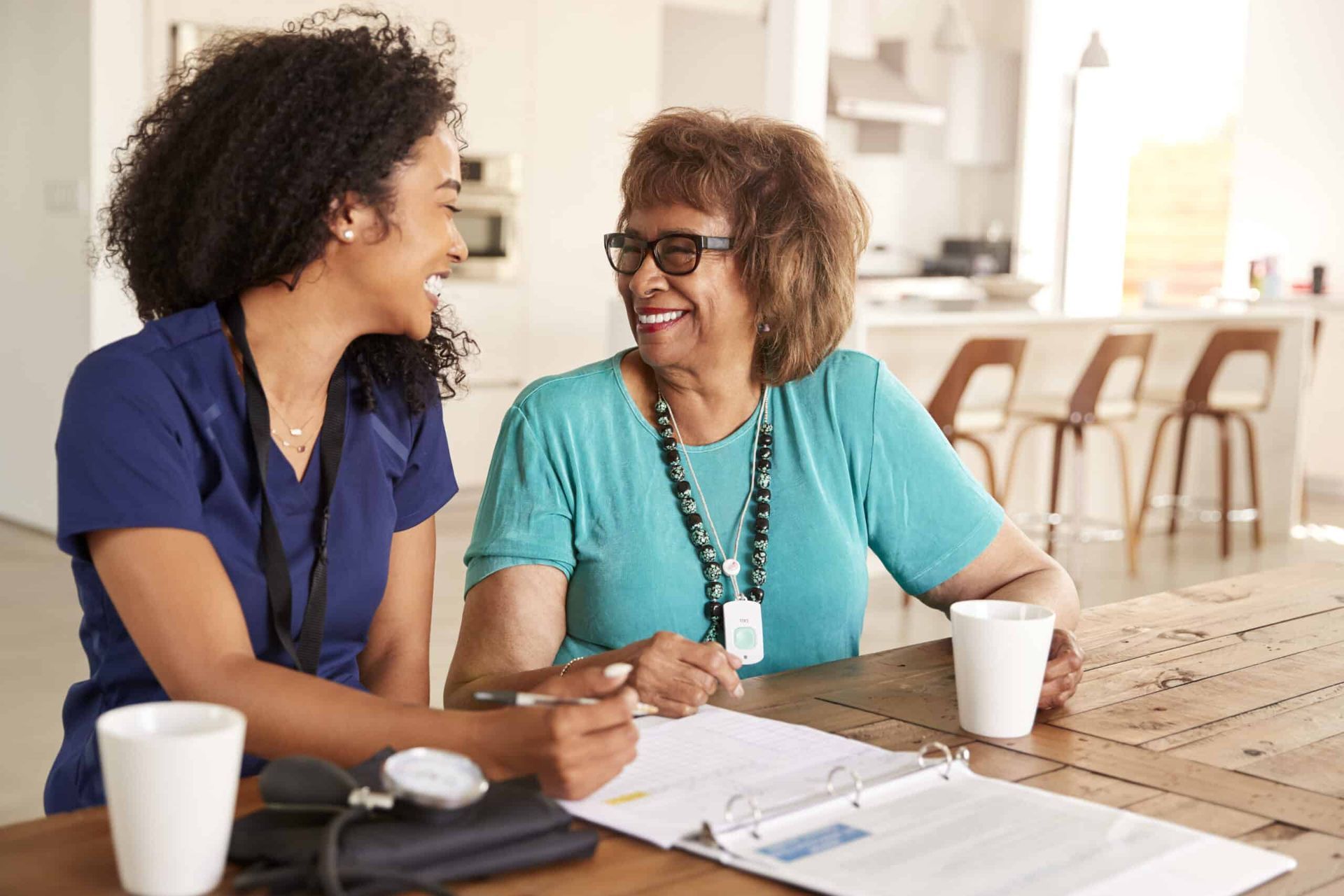 Caregiver and senior woman at table, smiling. Papers, blood pressure monitor, and coffee present.
