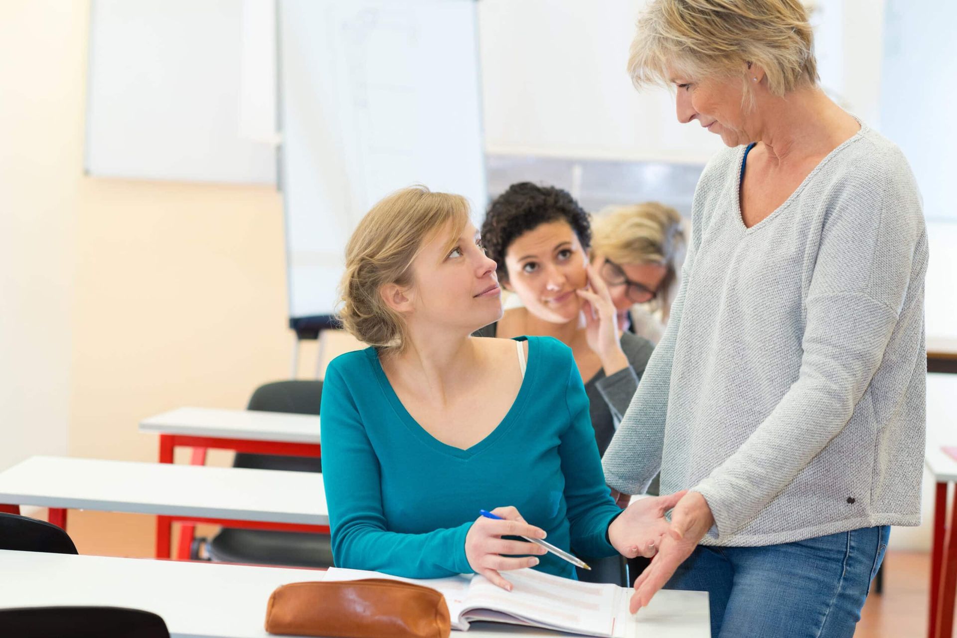 Two people talking in a classroom, with one seated at a desk holding papers and the other standing nearby.