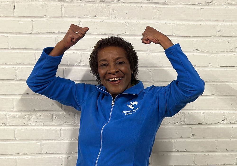 Woman in blue jacket, arms raised with fists clenched, smiling in front of white brick wall.