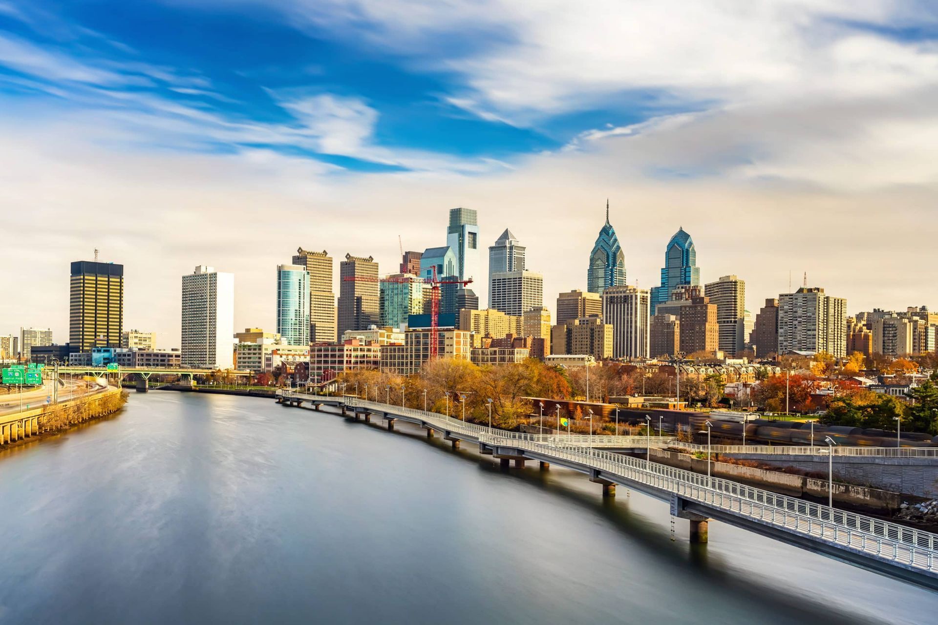 Philadelphia skyline viewed across a river with a bridge, under a blue sky.