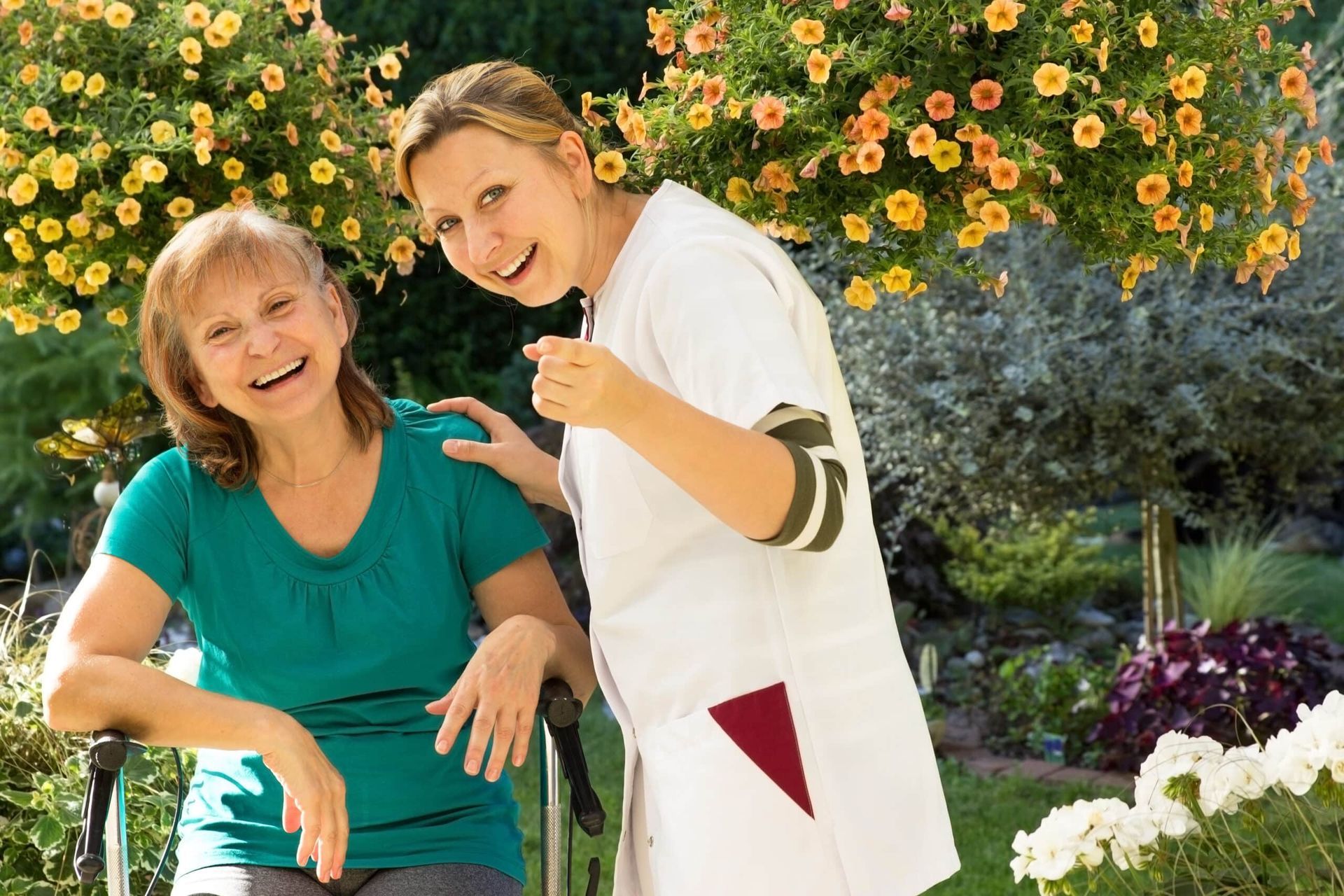 Woman in wheelchair laughs with a caregiver in a garden with flowers; both smile.