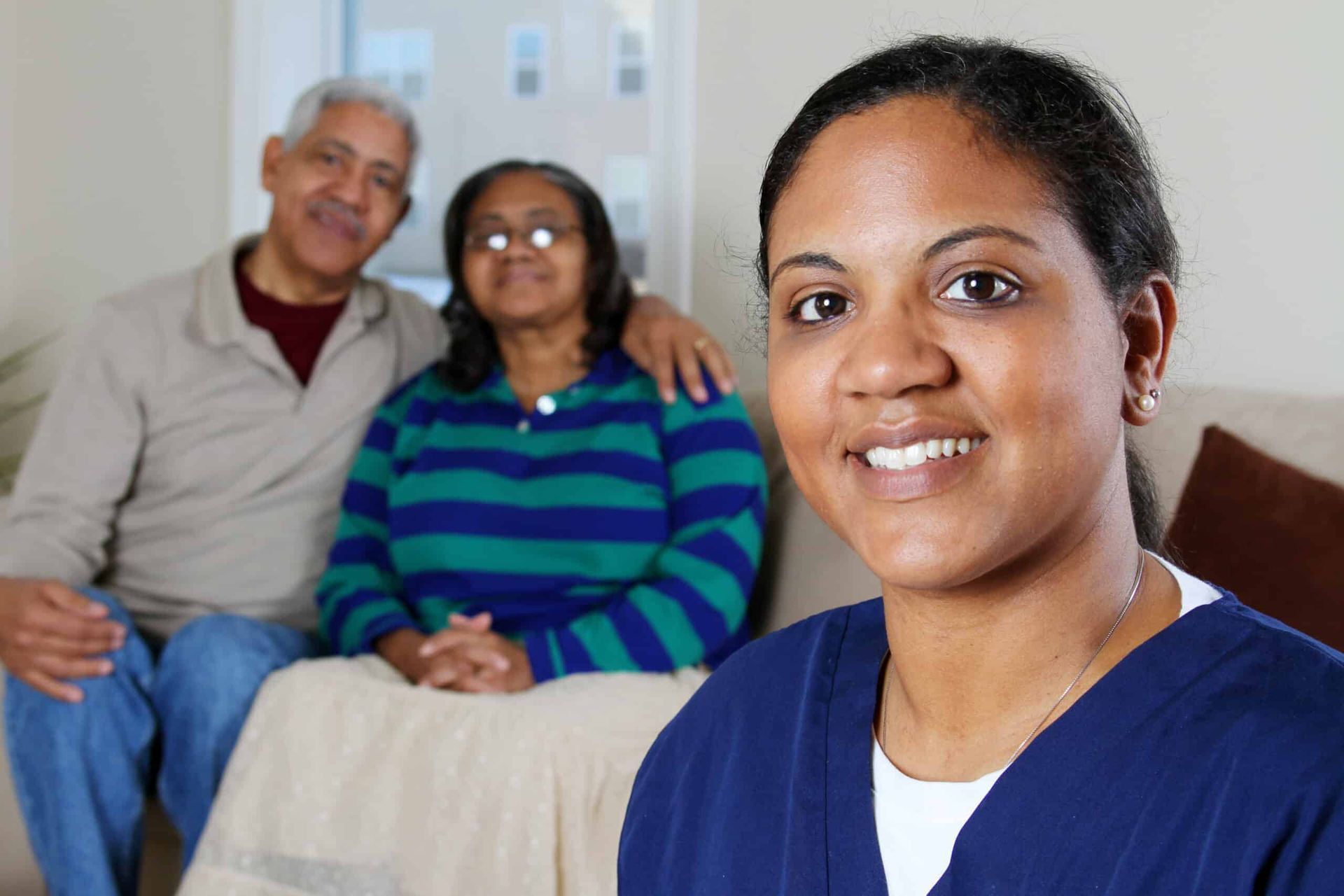 Caregiver smiling, with elderly couple on couch in the background.