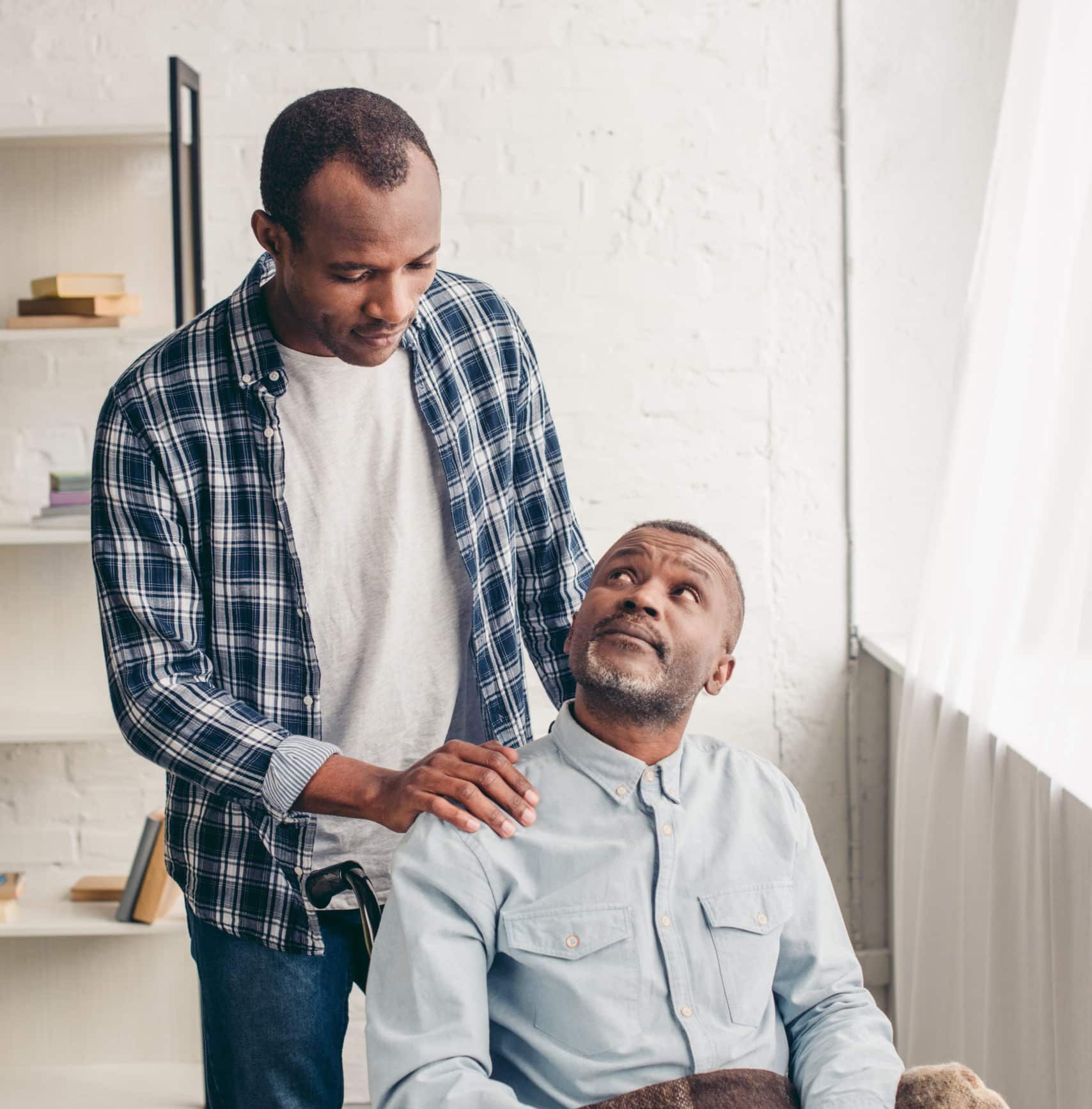 Man placing a hand on an older man's shoulder; both are African American. Interior setting.