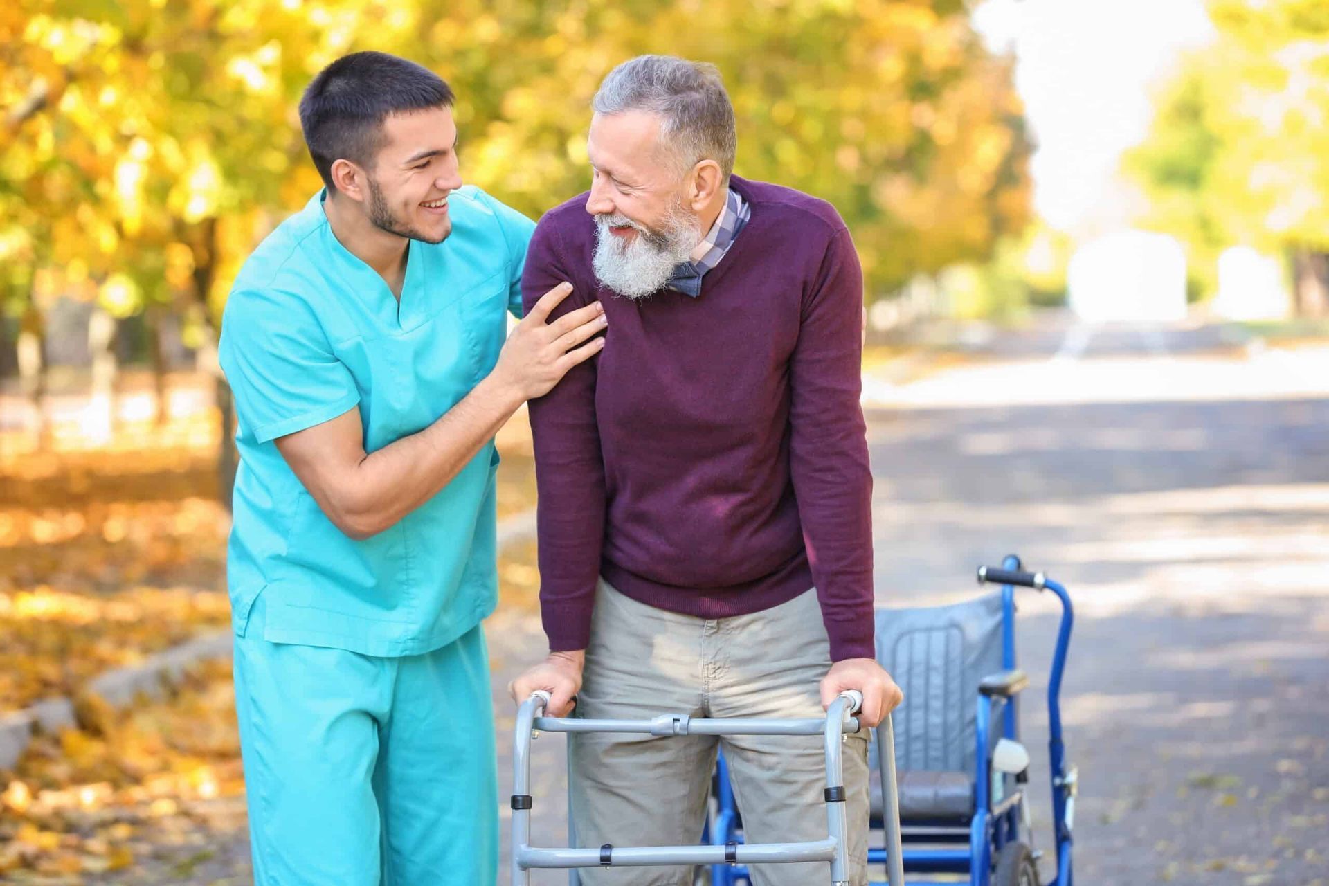 Caregiver assists elderly man using a walker outdoors on a sunny path.
