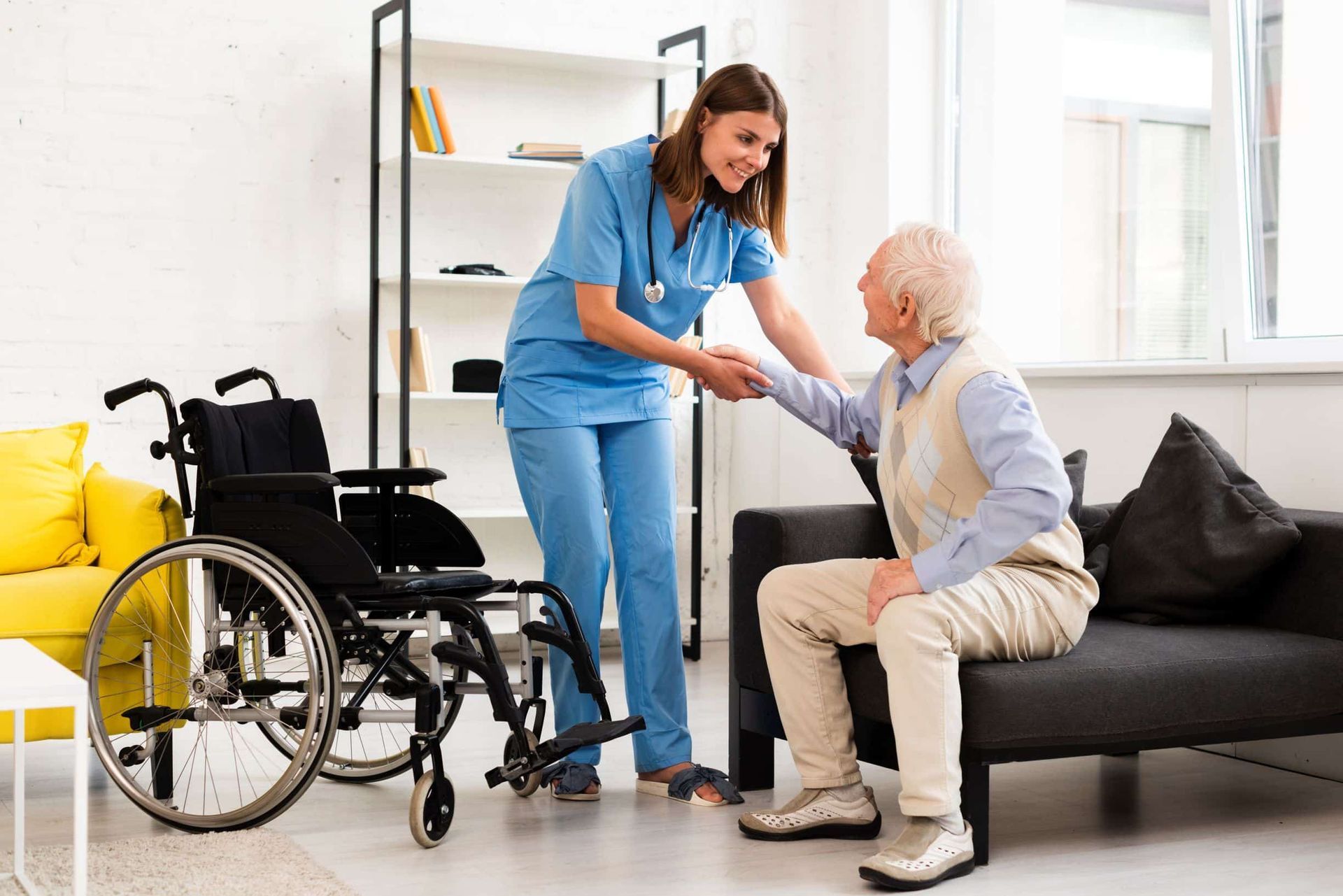 Nurse in blue scrubs assists elderly man from couch; wheelchair nearby.