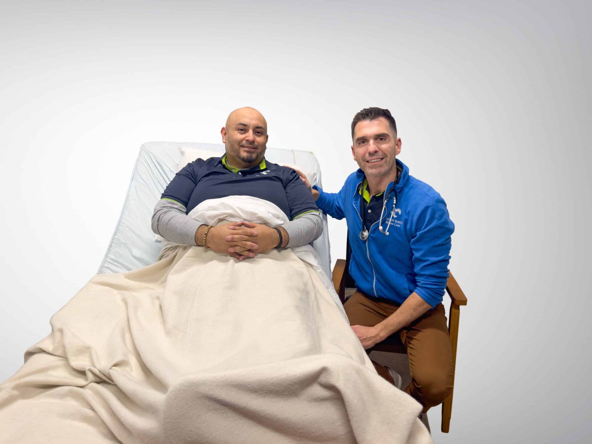 A man in bed smiles as a man in a blue jacket kneels beside him, hand on his shoulder.