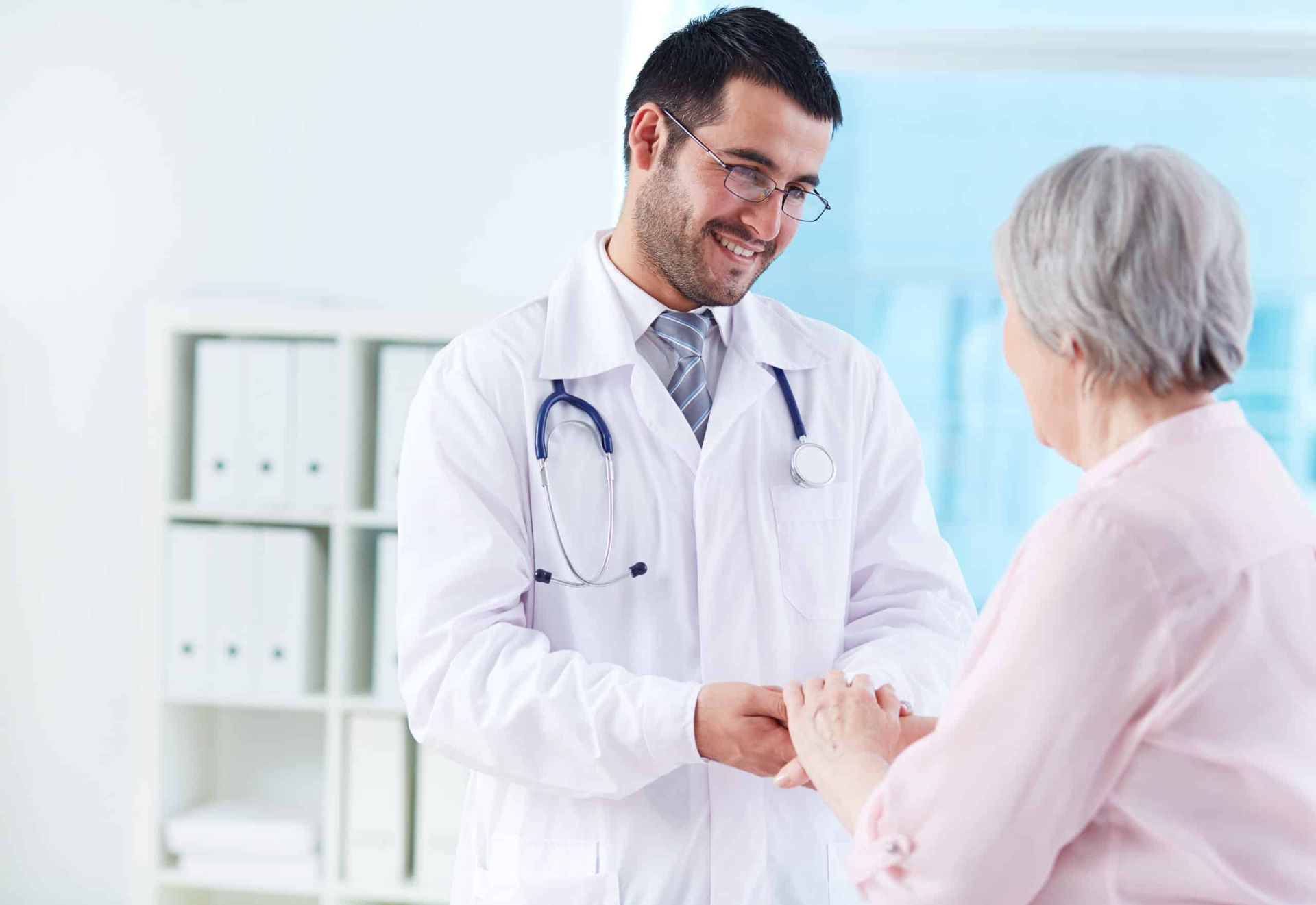 Doctor holding a patient's hand, smiling in a light-filled office. They are both white.