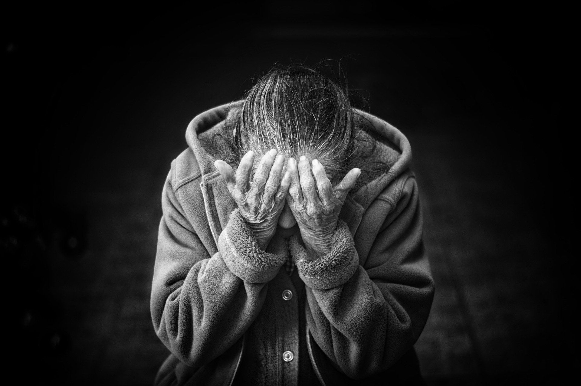 A person in a hooded coat and gloves sits hunched with face buried in their hands against a dark background.