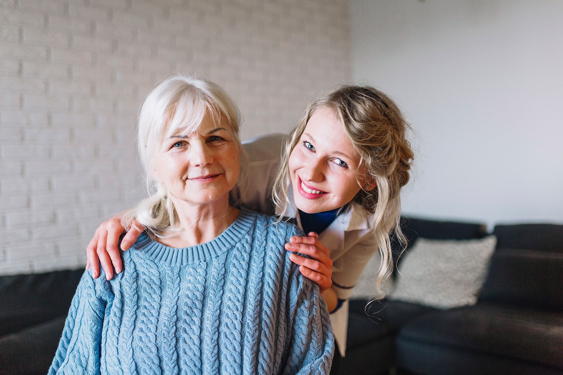 Woman with hands on shoulders of older woman; both smiling in a home.