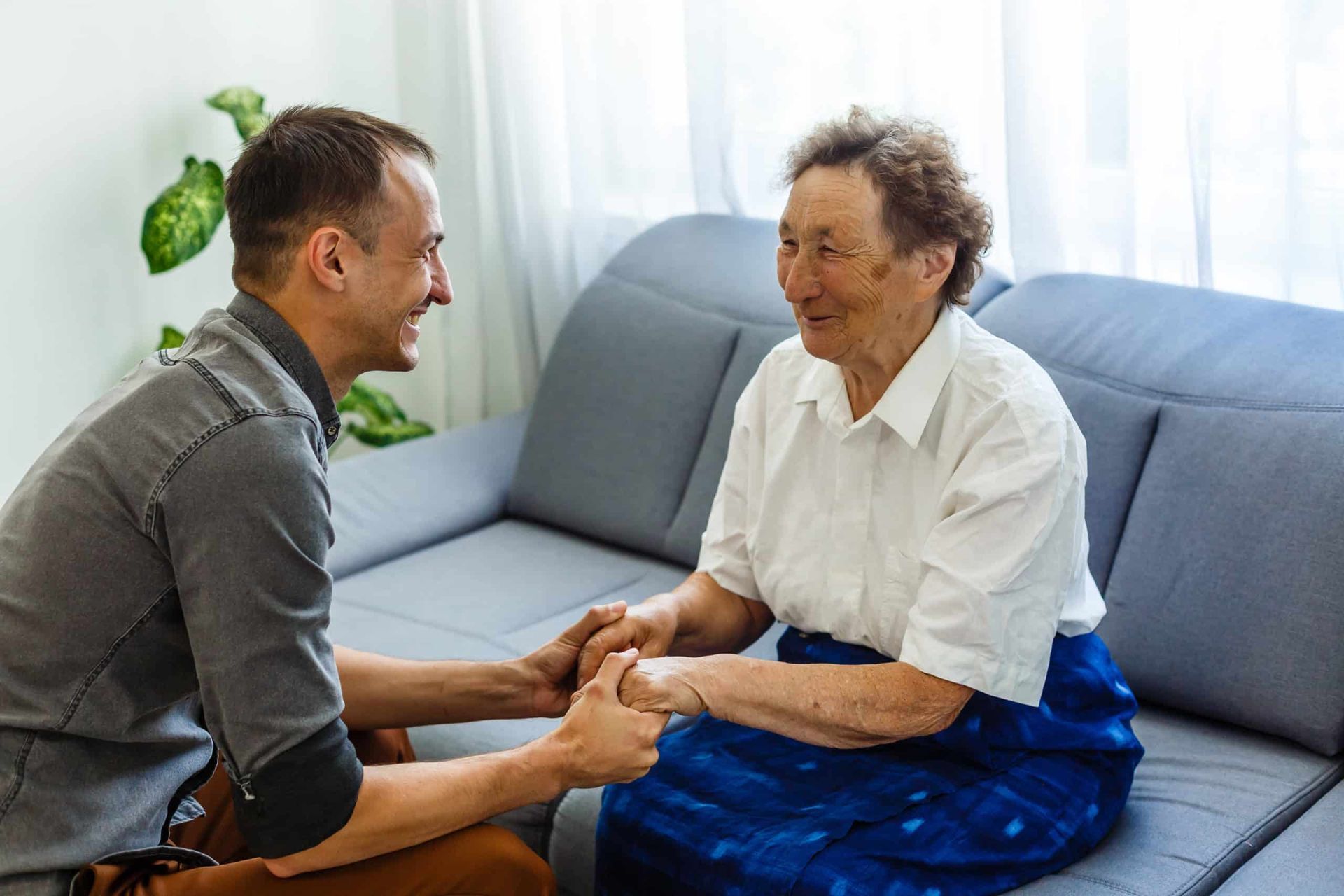 Man smiling, holding hands with elderly woman on a couch. They appear happy indoors, good lighting.