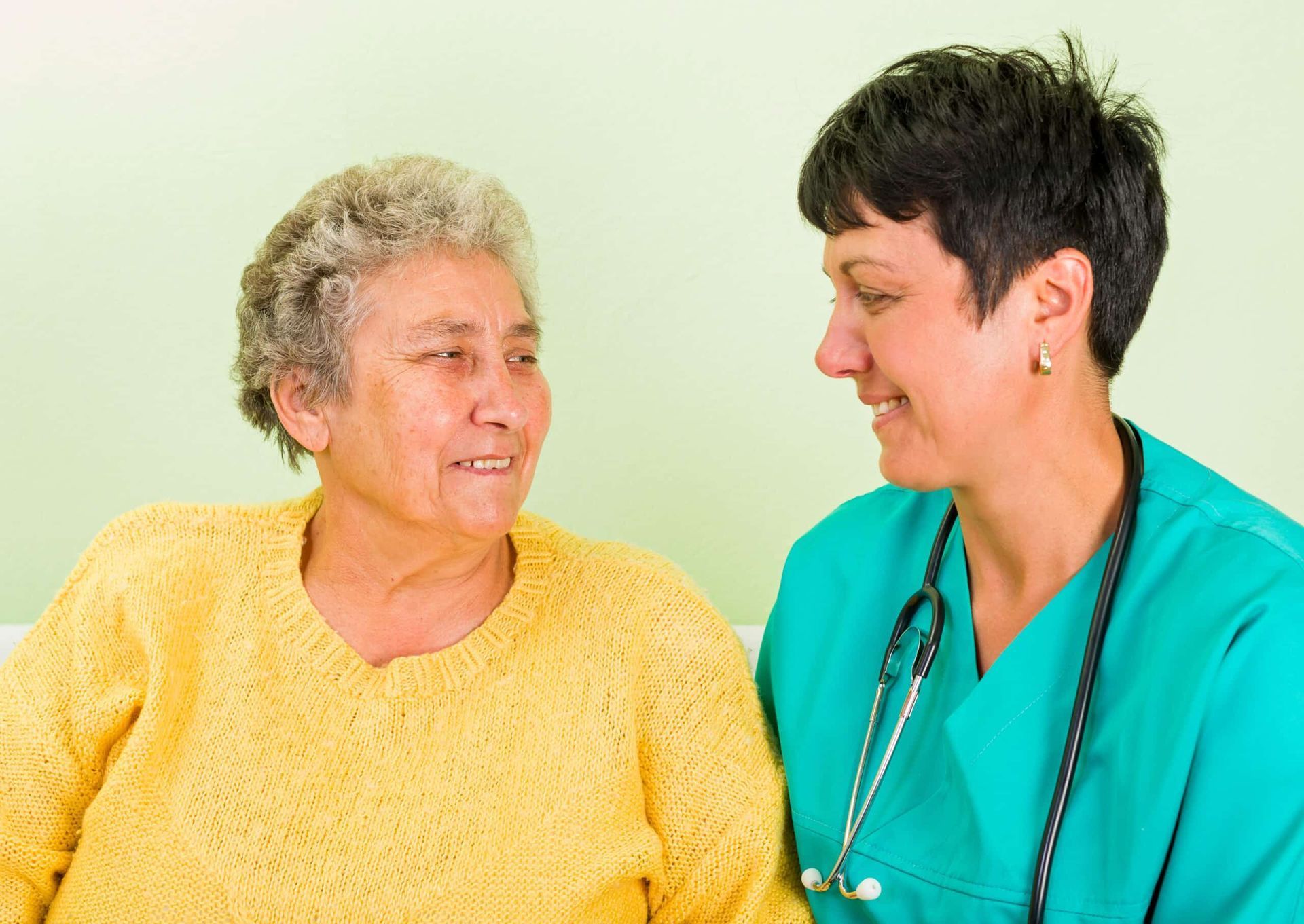 Woman in yellow sweater smiles at a medical professional in green scrubs, both looking at each other.