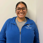 Smiling person in glasses and a blue zip-up jacket standing against a light wall