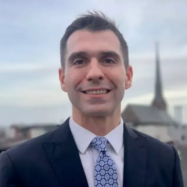 Smiling man in a dark suit and patterned tie, standing outdoors with the Eiffel Tower in the background