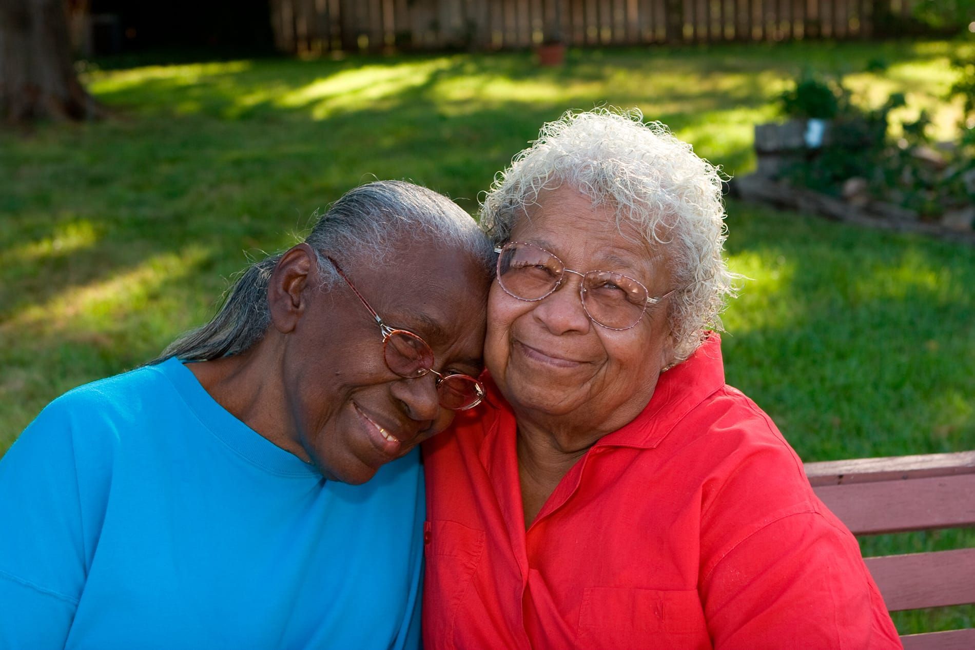 Two elderly Black women smiling and embracing on a park bench. One wears a blue shirt, the other red.