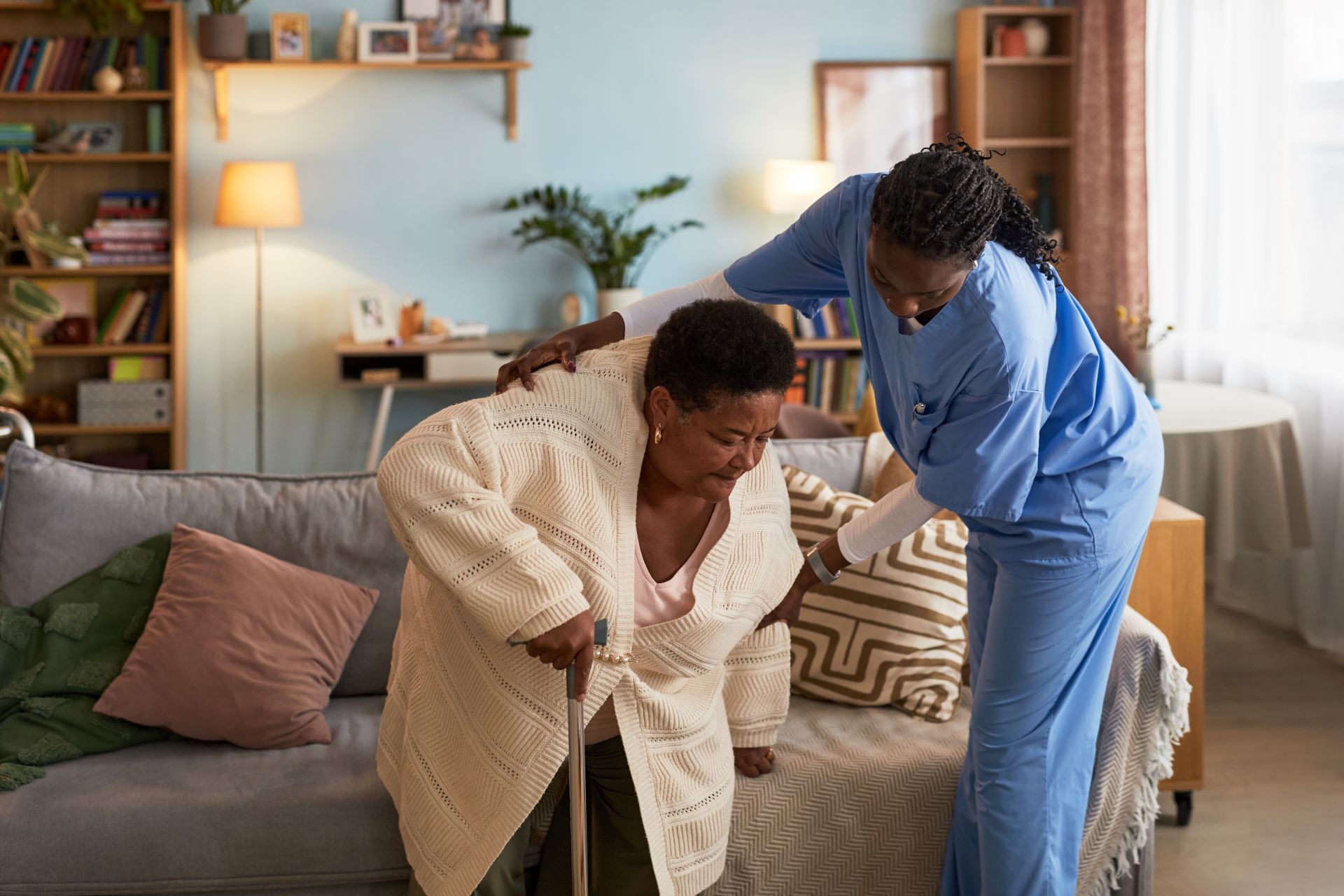 Two people helping someone stand in a cozy living room with a gray sofa and shelves.