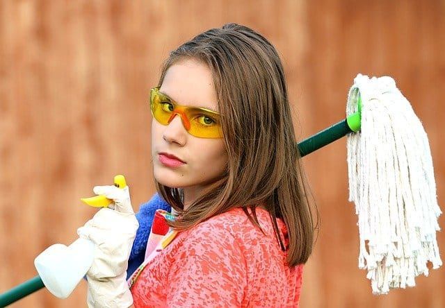 Young woman with safety glasses, holding a mop and spray bottle, looking stern.