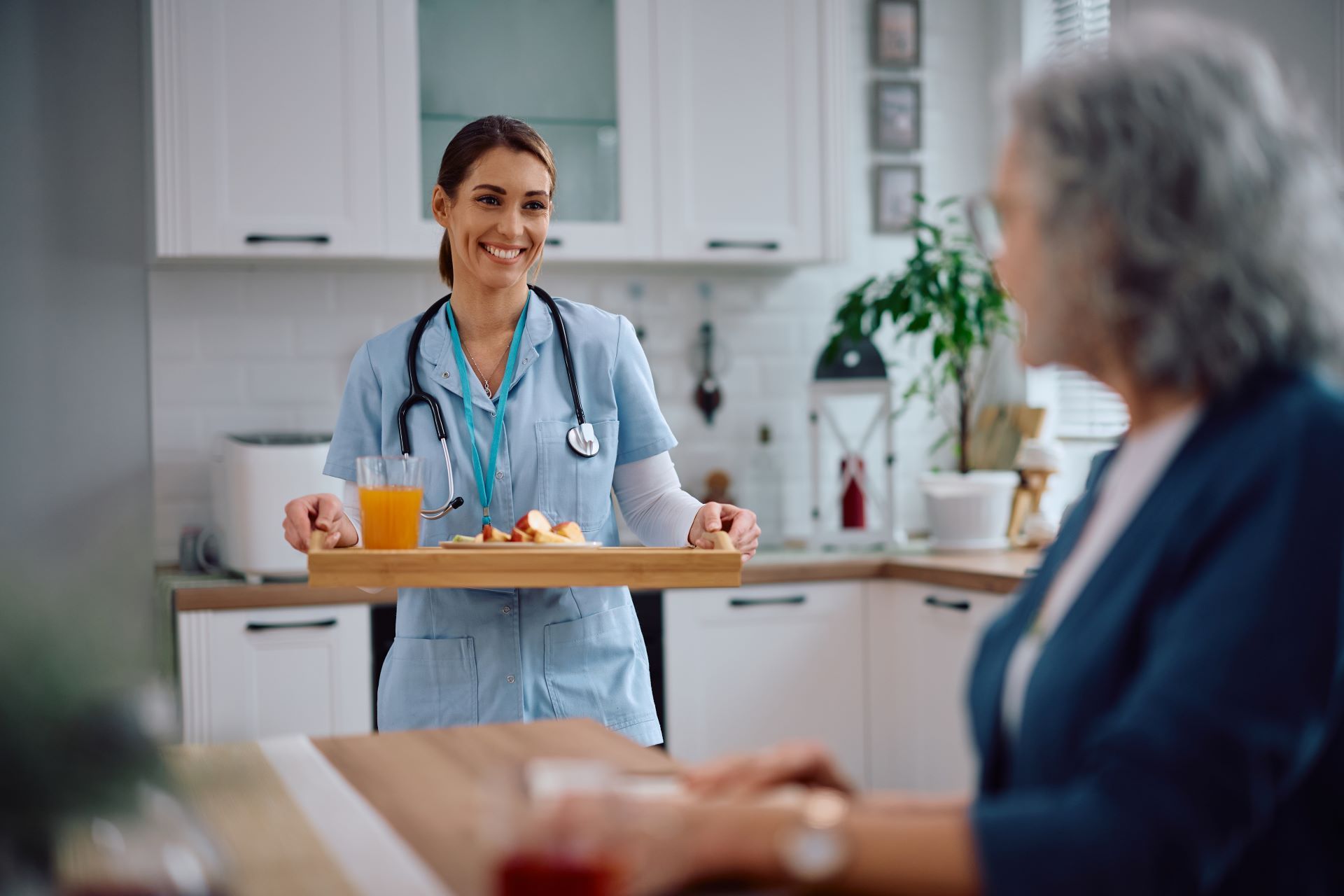 Smiling nurse serving breakfast on a tray to a seated patient in a bright kitchen-like room