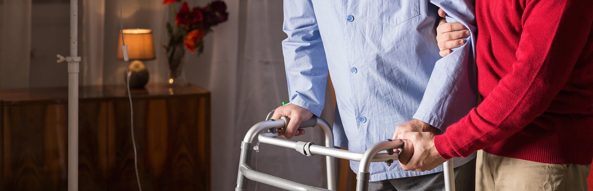 An older man using a walker is assisted by another person, indoors.