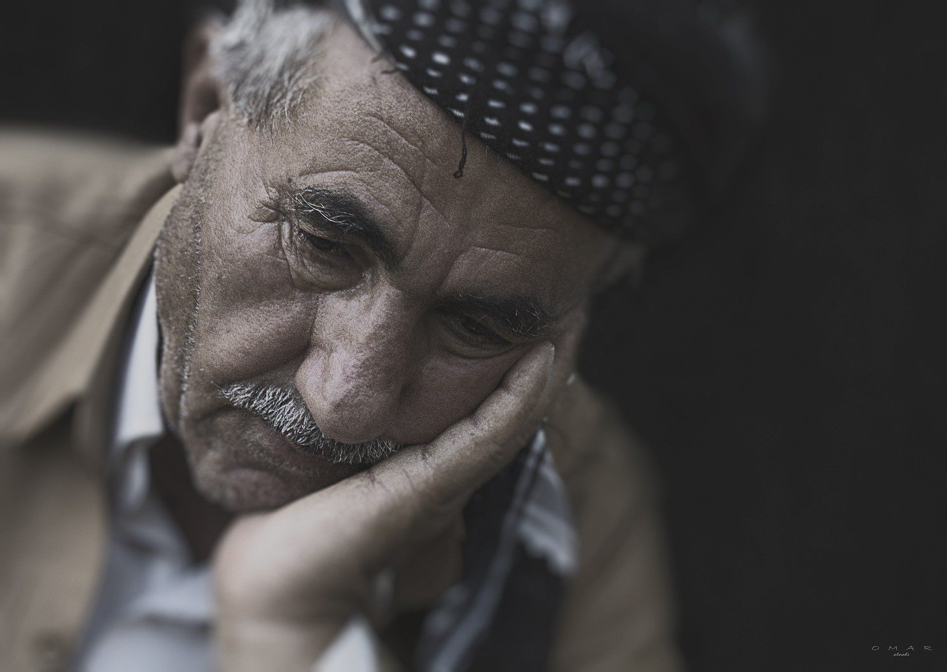Older man rests his head in his hand, looking down. He wears a dark cap and light brown jacket.
