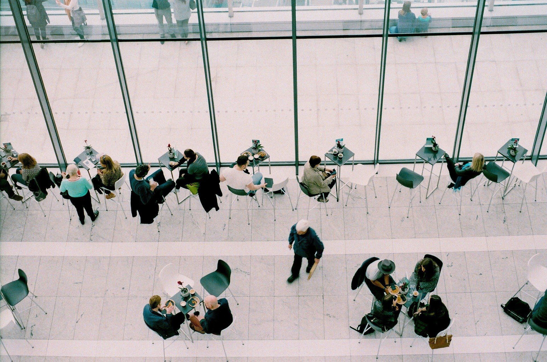 People in a bright lobby sit and stand by a glass wall, seen from above.
