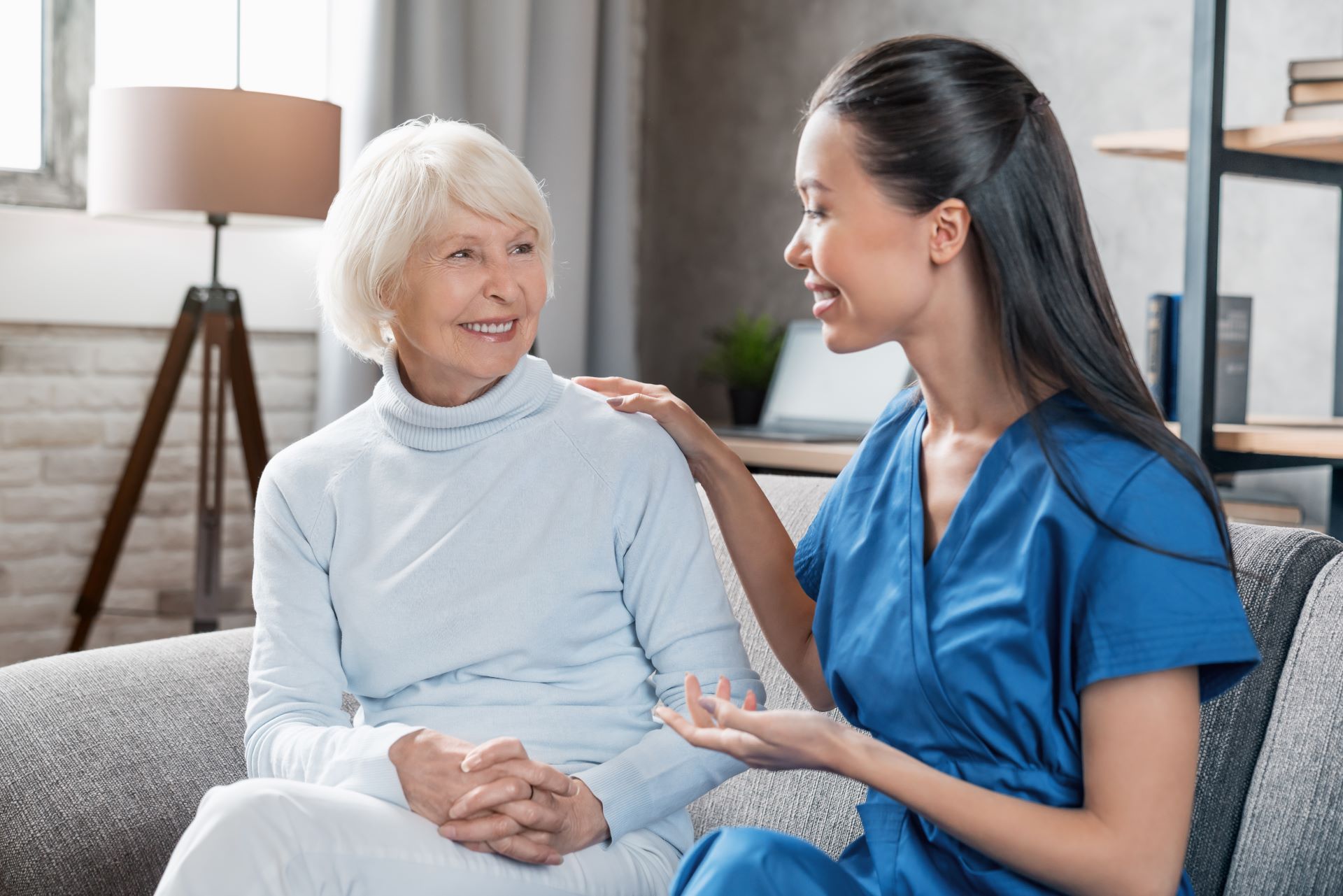 Two women talking on a couch, one in blue scrubs comforting an older woman in a light sweater.