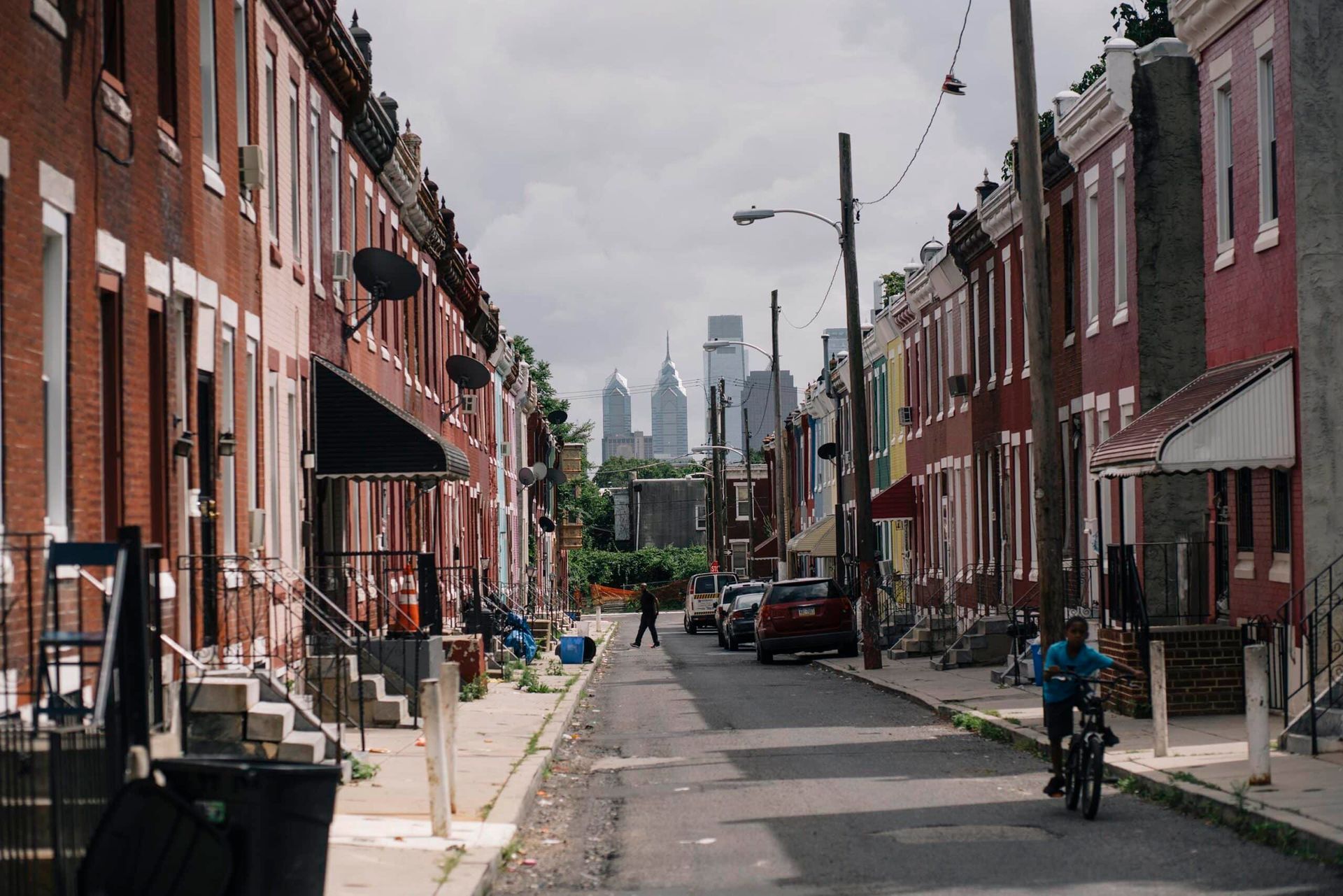 Row houses line a street; a child bikes towards distant skyscrapers under a cloudy sky.