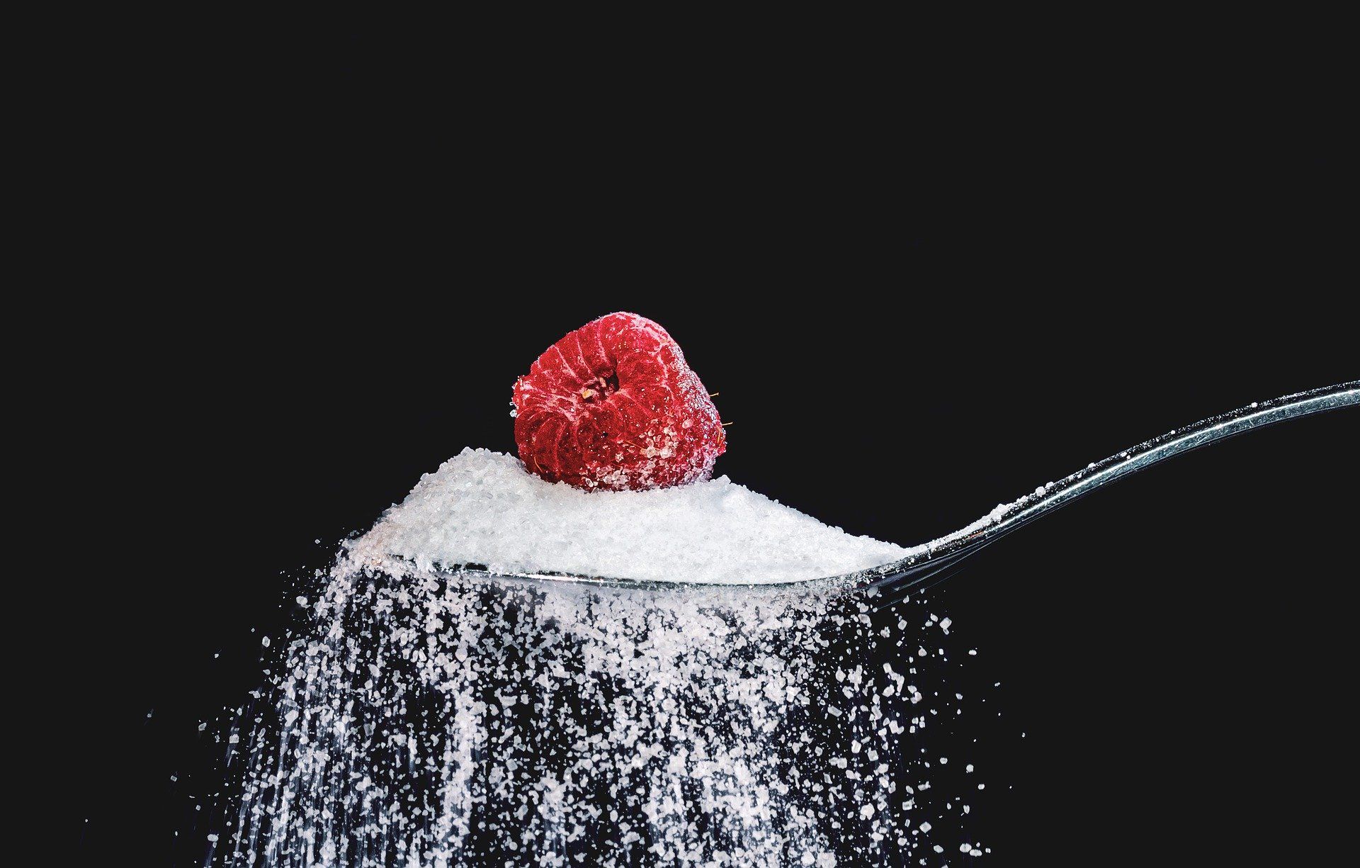A raspberry sits atop a spoonful of sugar, with sugar spilling out against a black background.