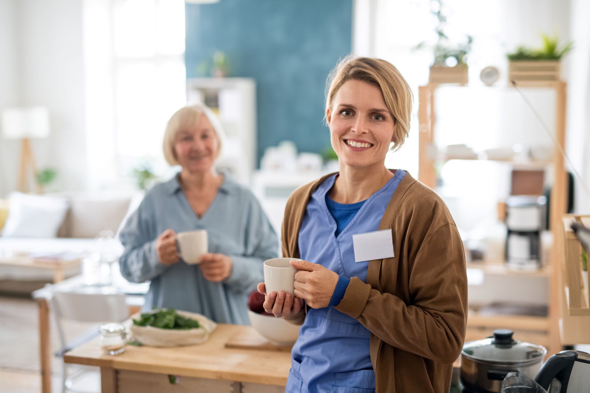 Two women holding mugs in a bright kitchen, smiling near a counter with food and pots