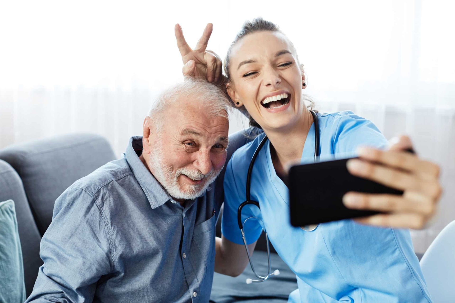 Caregiver and elderly man smiling, taking a selfie indoors; caregiver is giving peace sign.