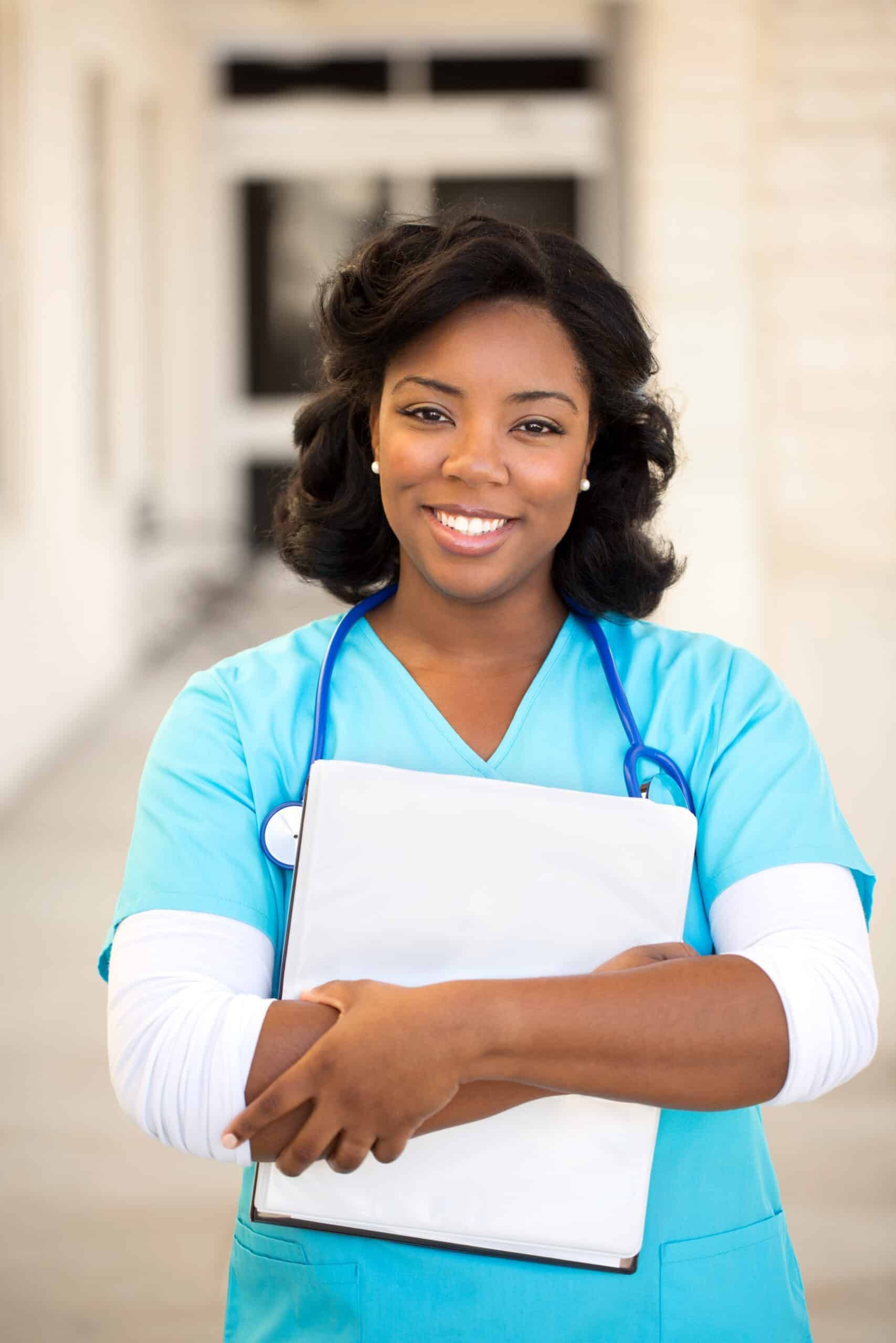 Smiling Black nurse holding a clipboard, wearing blue scrubs and a stethoscope outside a building.