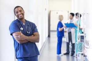 Smiling healthcare worker in blue scrubs leans against a wall in a hospital hallway; colleagues in the background.