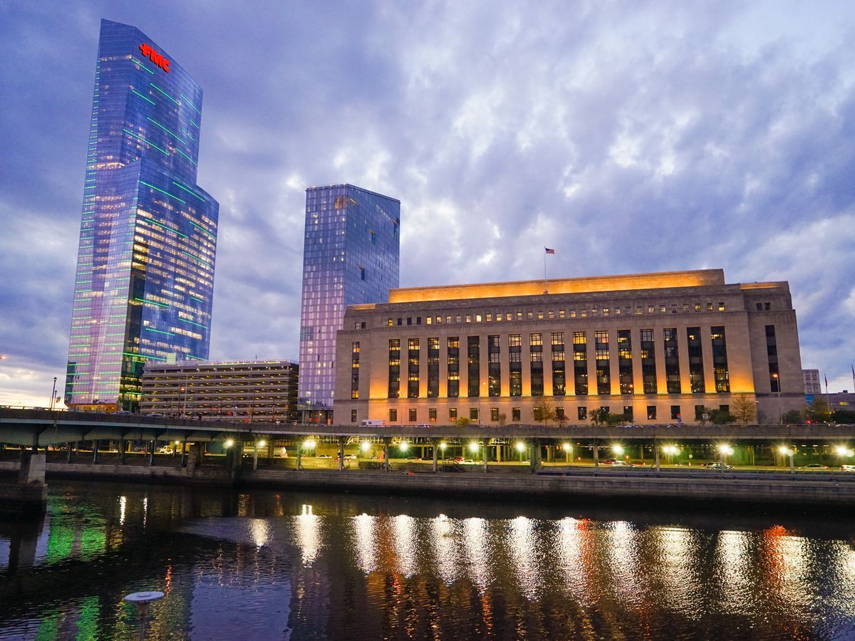 Philadelphia skyline at dusk; high-rise buildings reflect lights on water, under a cloudy sky.