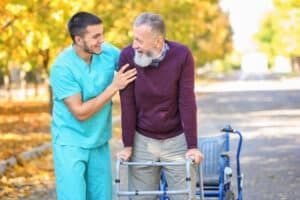 Man in scrubs assisting elderly man using a walker outdoors, smiling.