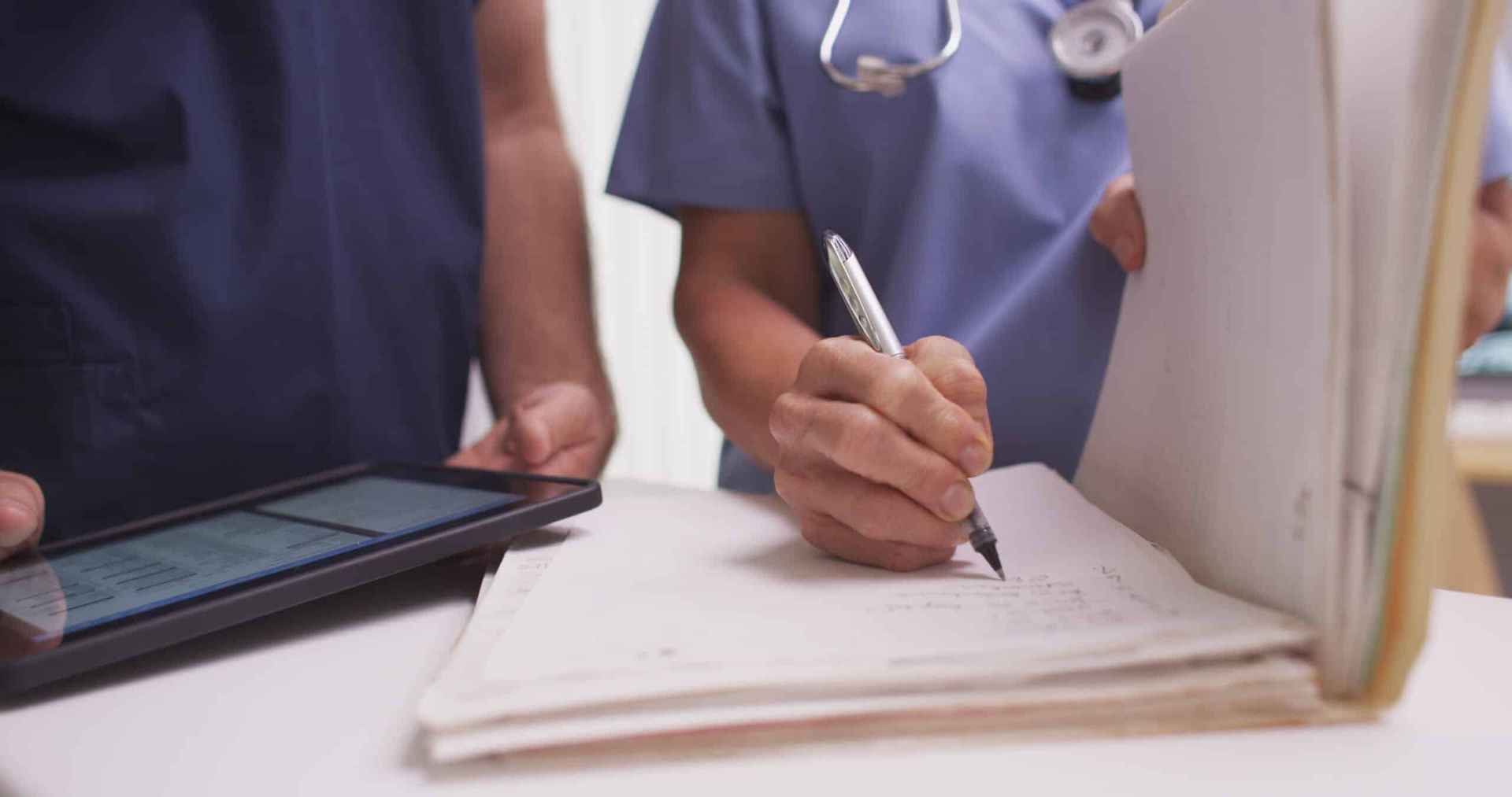 Two medical professionals reviewing patient records. One writes with a pen; the other holds a tablet.