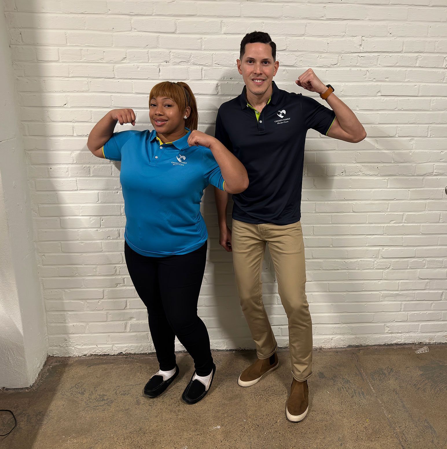 Two people flexing their biceps, smiling in front of a brick wall.