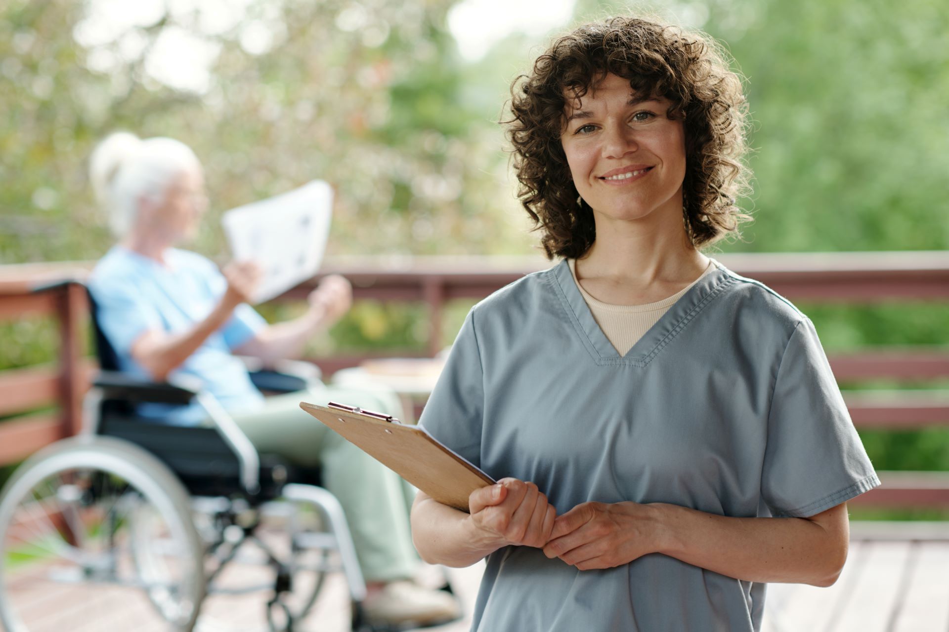 Healthcare worker in gray scrubs holding a clipboard, with a wheelchair user reading in the background.