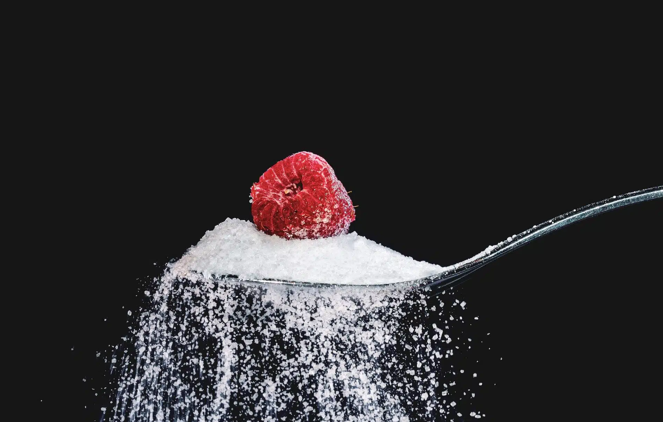 Raspberry on spoon piled high with sugar, some sugar spilling; black background.